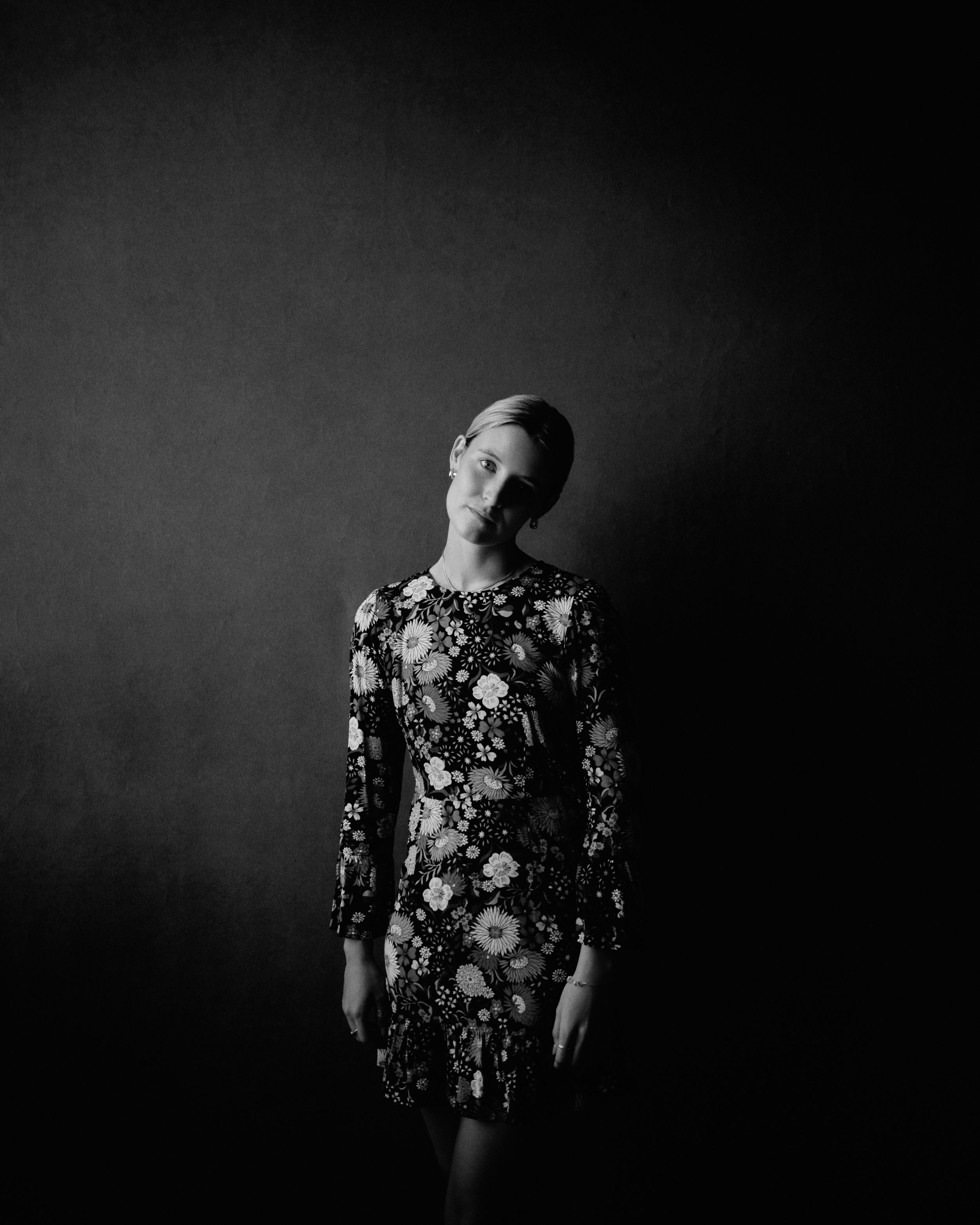 A black and white photo of a woman with short hair, wearing a floral dress, standing against a plain dark background with a slight tilt of her head.