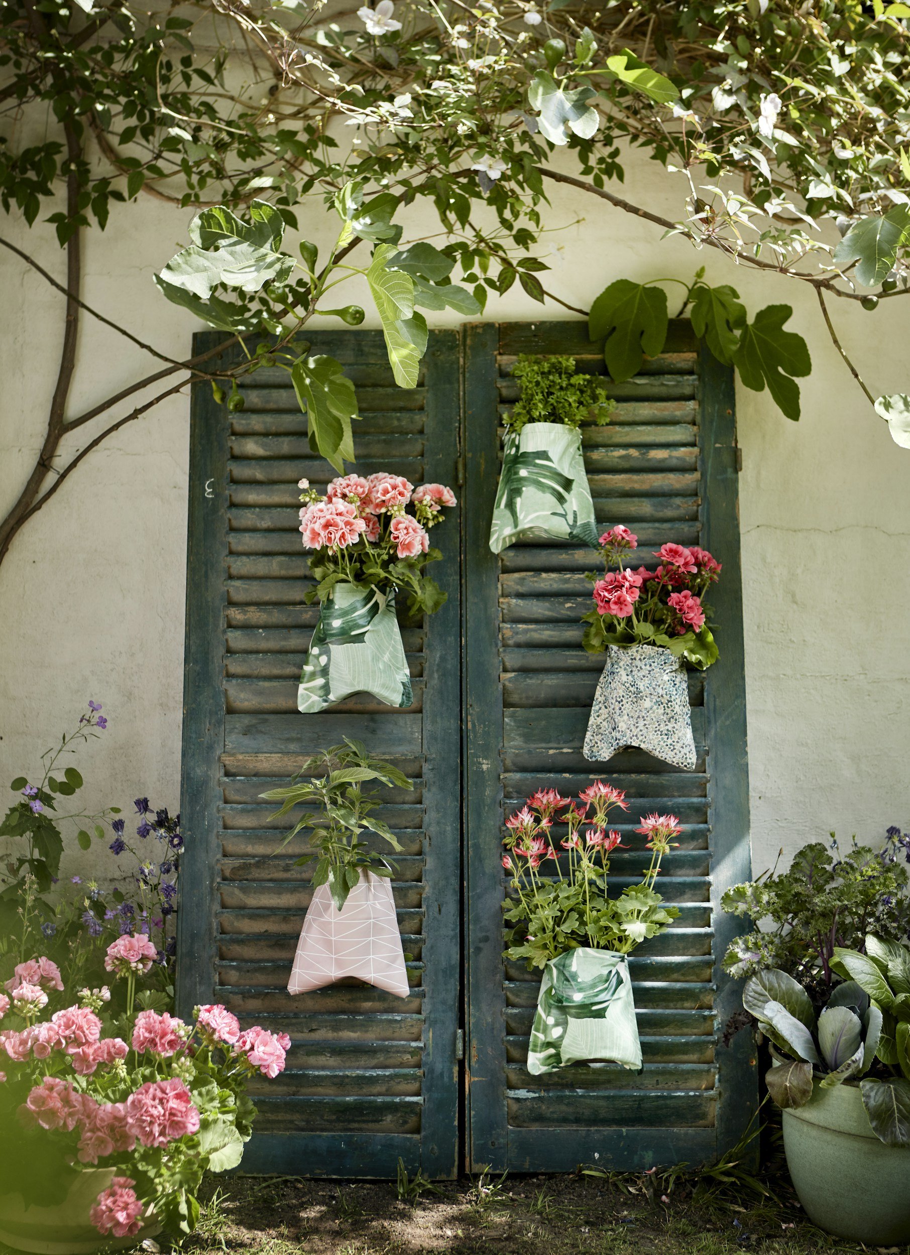 Old wooden shutter doors with hanging fabric bags filled with green plants, surrounded by colorful flowers and lush greenery.