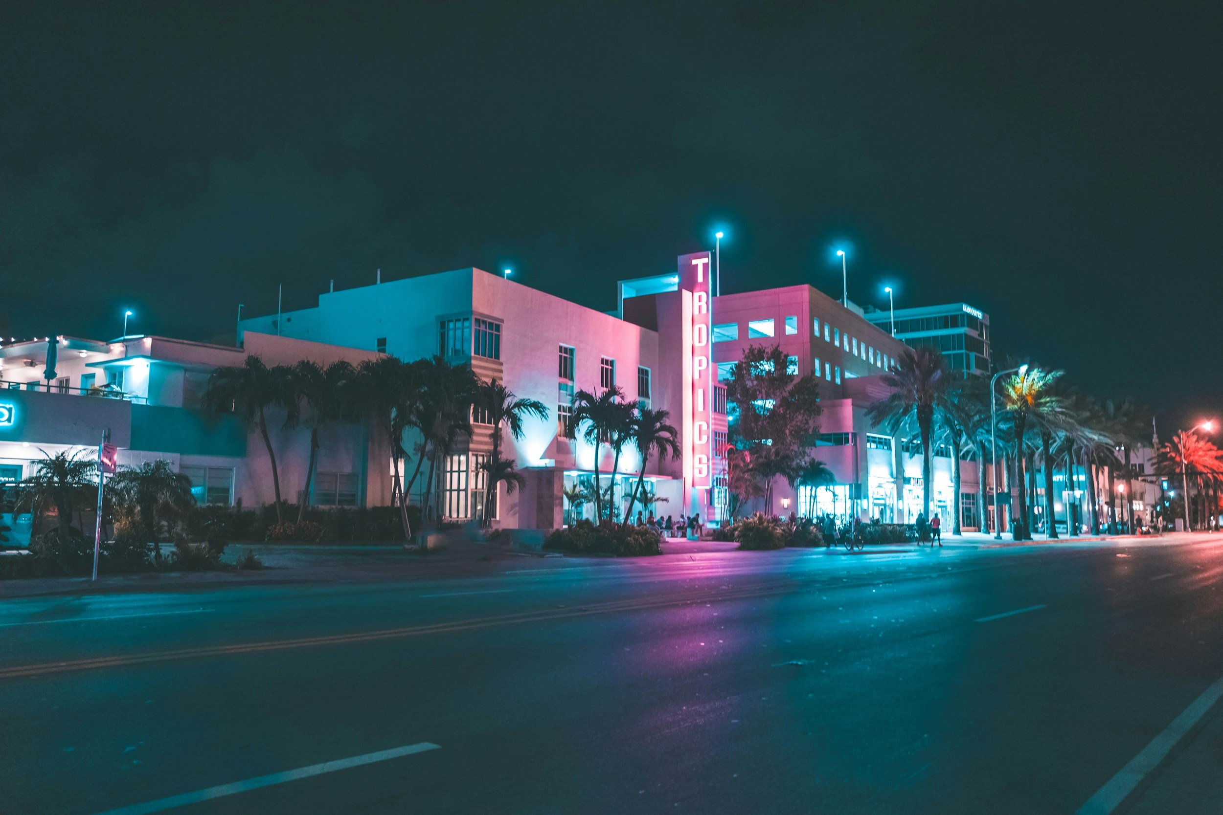 Night view of an art deco building with neon pink and blue lights, with palm trees in front and a sign that reads 'TR0OPICS'.
