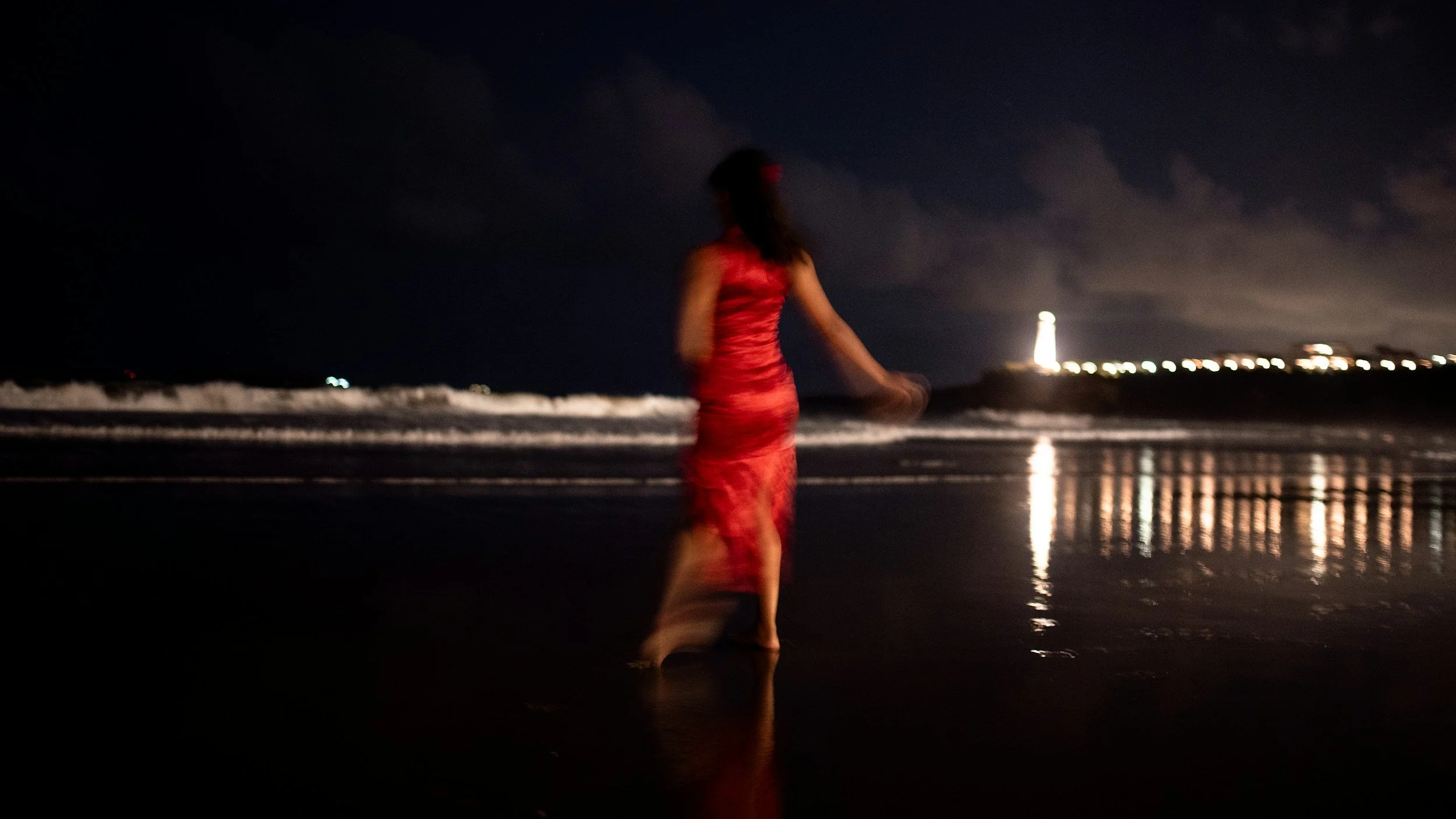 A woman in a red dress walking on the beach at night with a lighthouse and city lights in the background.