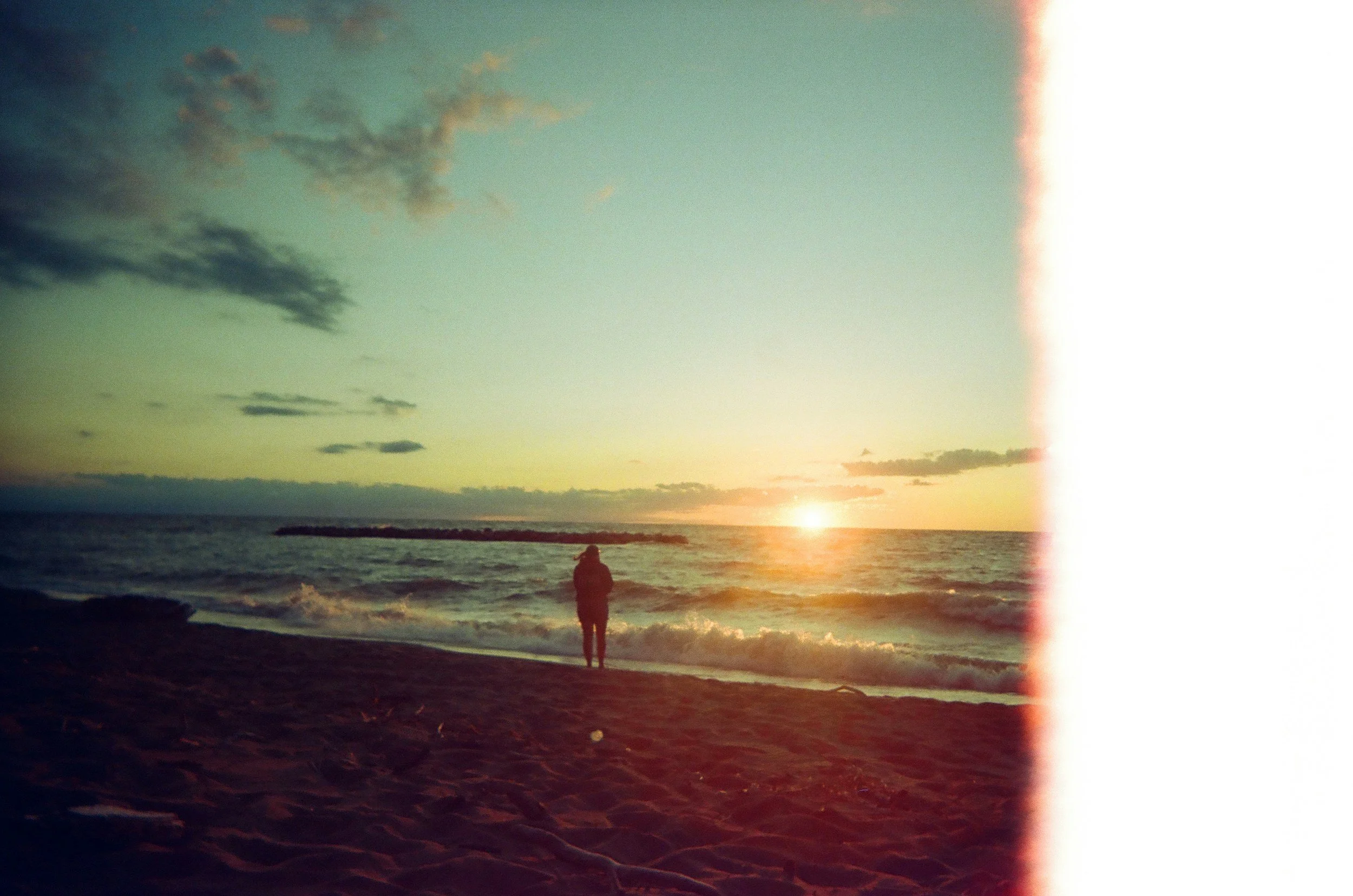 A person standing on the beach at sunset, with the ocean waves and cloudy sky in the background. The right side of the image appears overexposed or damaged, creating a light streak.