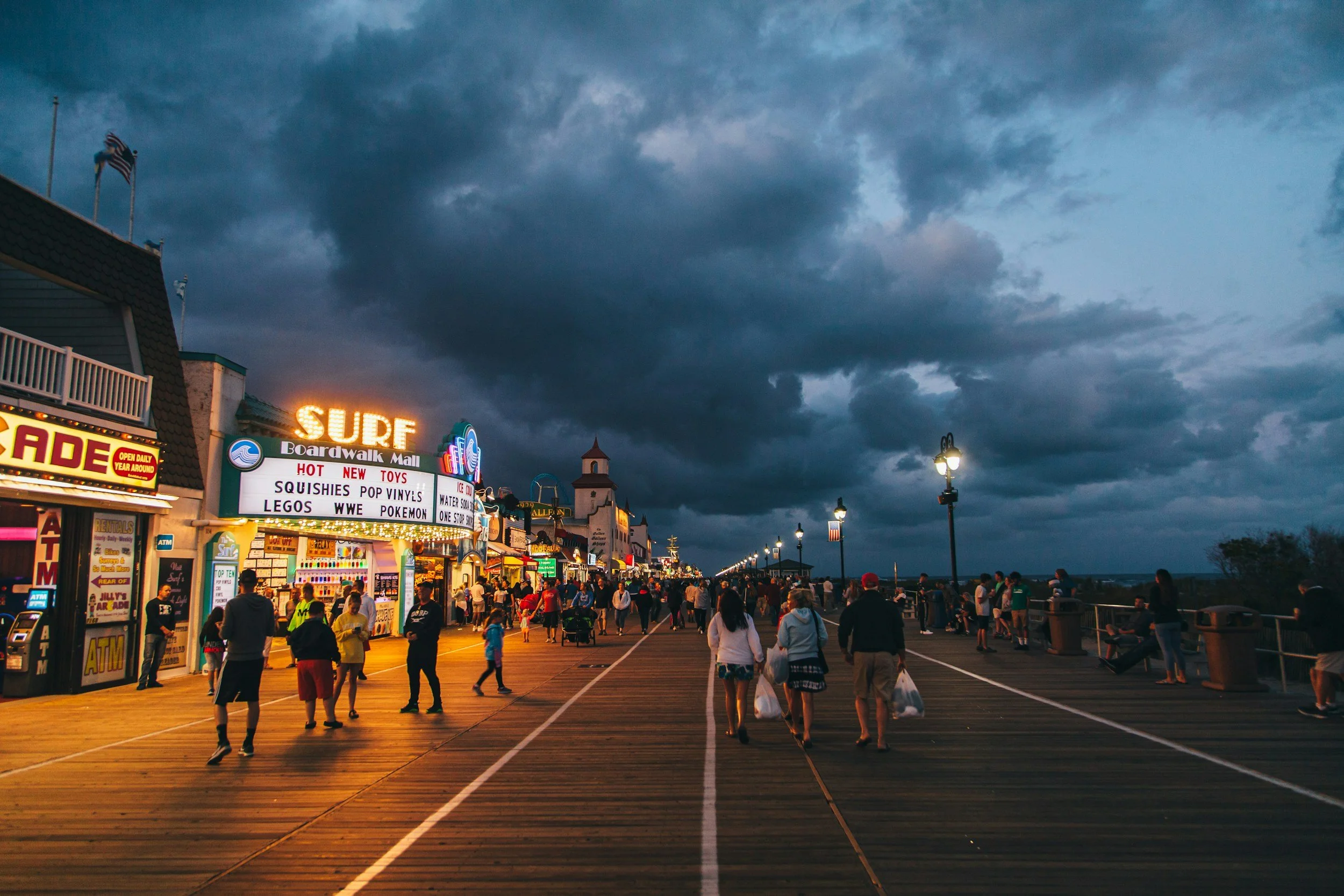 People walking along a busy boardwalk under a dark, cloudy sky at dusk, with shops and amusement signs illuminated.