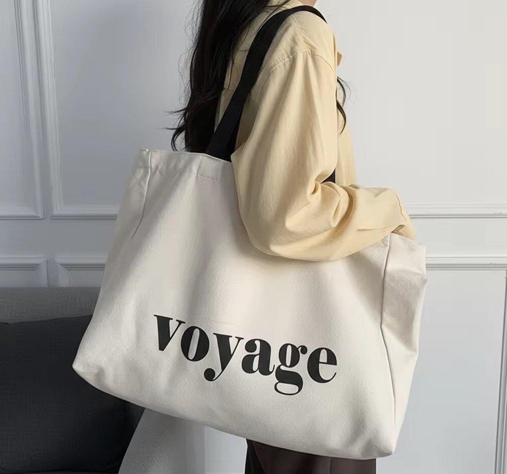 Person with long dark hair wearing a beige shirt carrying a large white tote bag with the word 'voyage' printed on it in black letters.