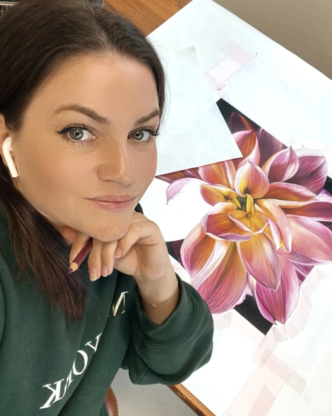 A woman with blue eyes and brown hair, wearing a dark green hoodie, is sitting at a table with a large, detailed flower illustration in front of her. She is smiling slightly and resting her chin on her hand, with white earbuds in her ears.