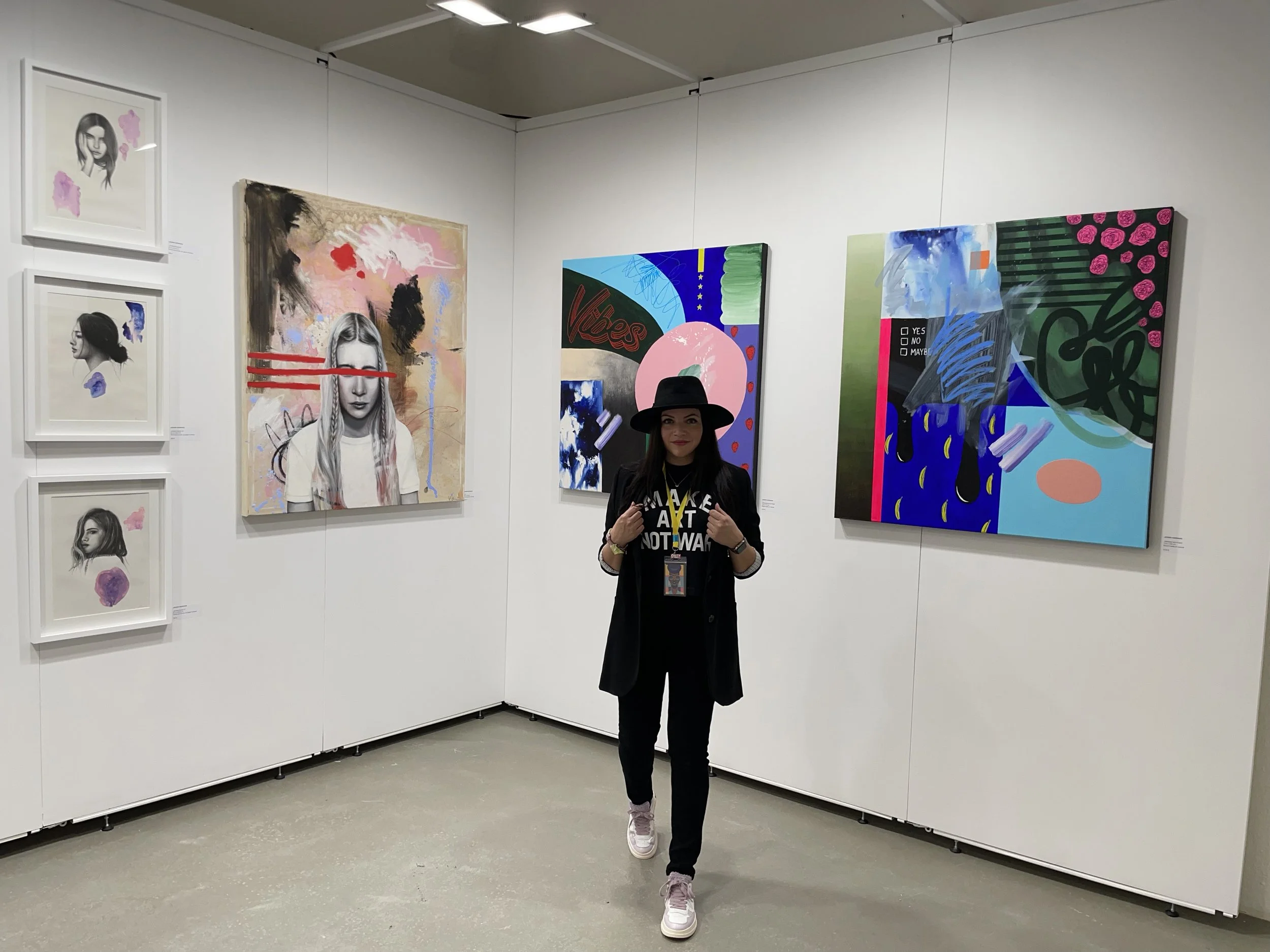 Woman wearing a black hat, black blazer, and sneakers standing in an art gallery with colorful paintings on white walls.