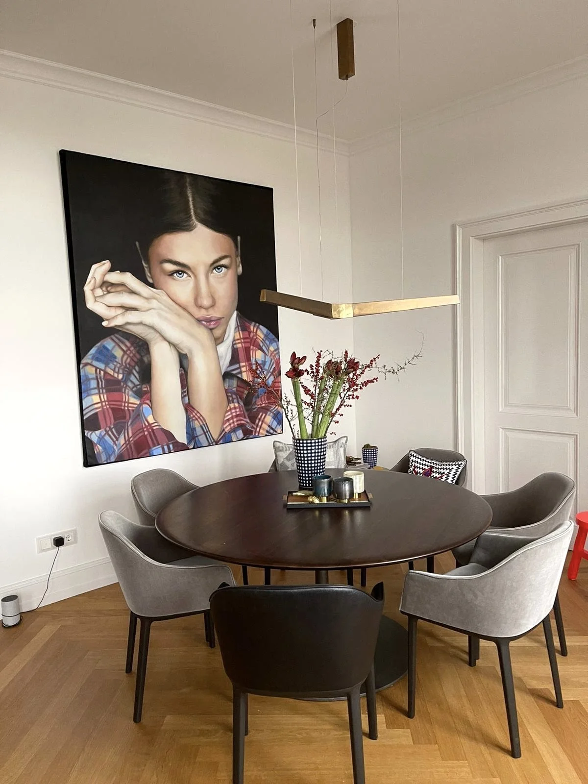 Dining room with a large portrait of a woman in a checkered shirt hanging on the wall, a round wooden table with a floral centerpiece, and modern chairs surrounding the table.