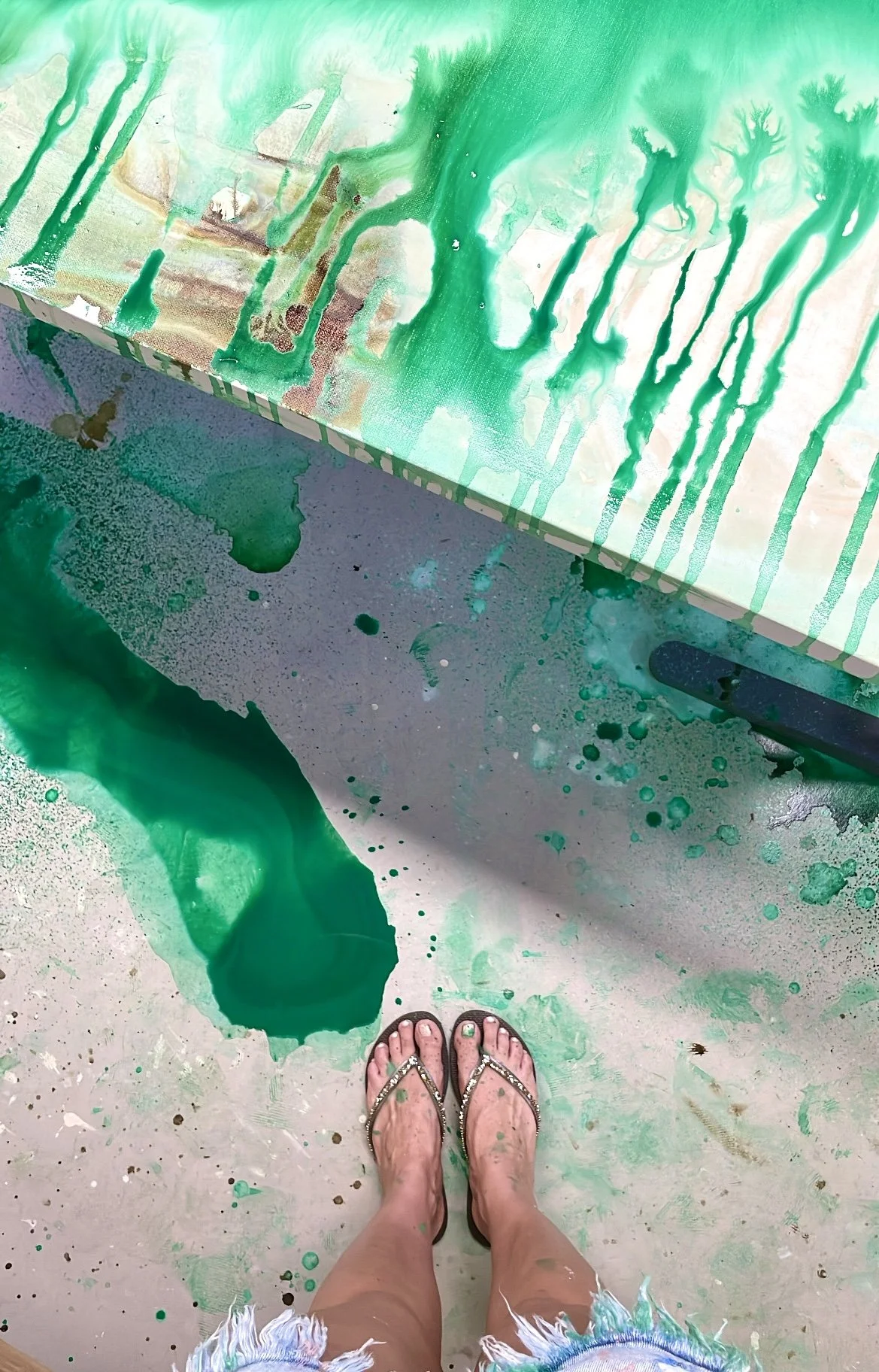 Person standing in front of a colorful abstract painting with green and blue paint splatters on the floor and canvas, wearing tan sandals and frayed denim shorts.