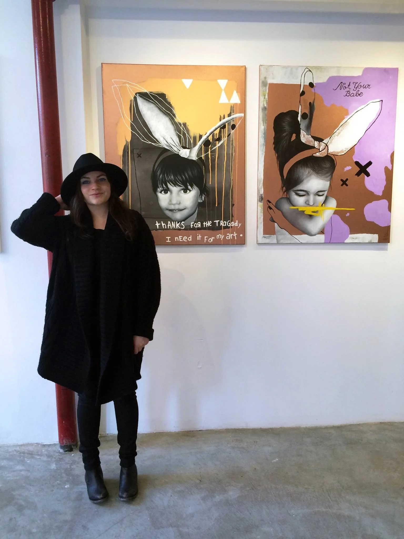 A woman with long dark hair wearing a black coat and wide-brimmed black hat standing in front of two abstract art pieces with black and white portraits of girls,one with a bunny ear and the other with a bow and a yellow airplane nose