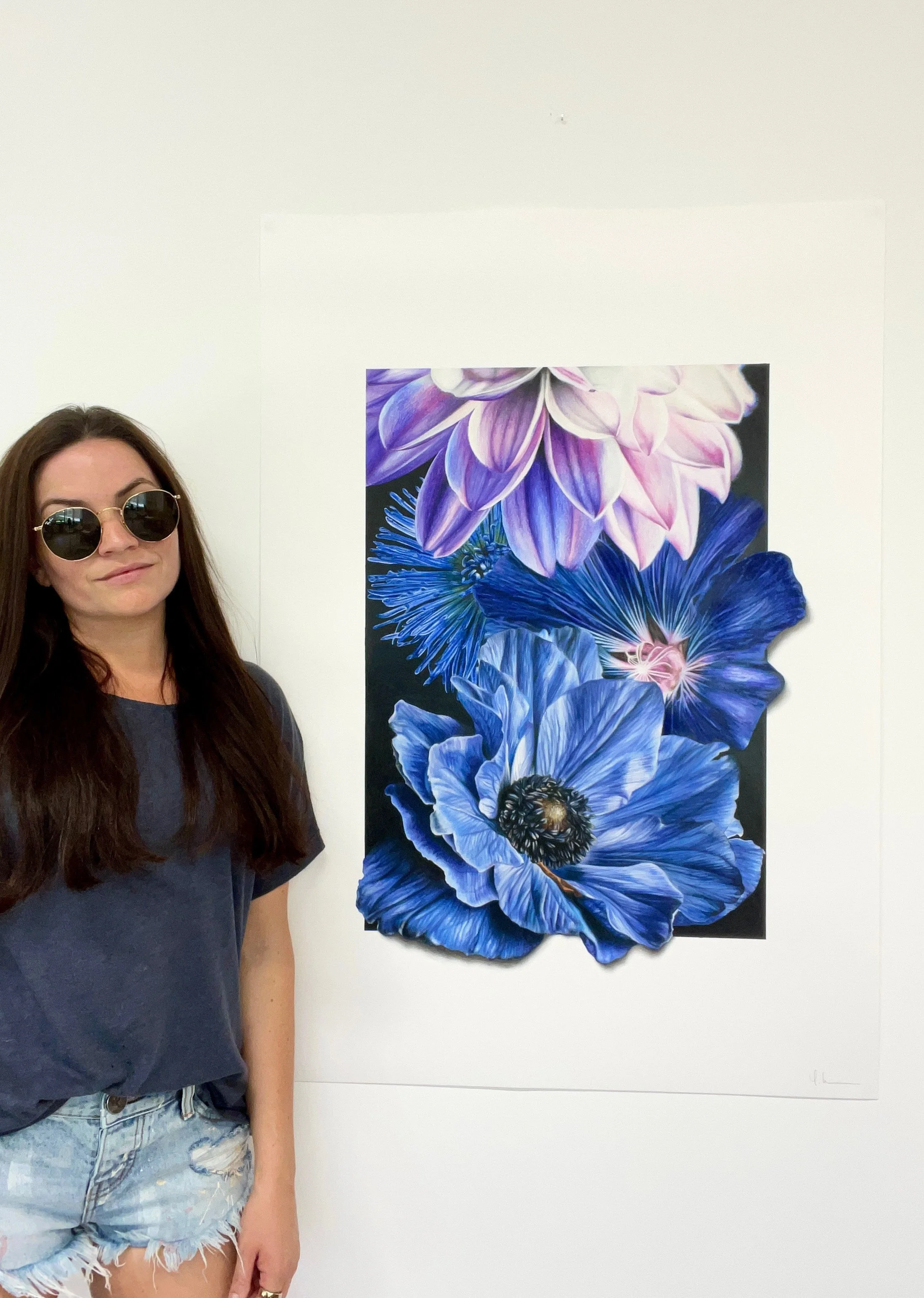 A woman with long dark hair wearing sunglasses and a loose gray t-shirt, standing next to a large colorful flower painting on a white wall.