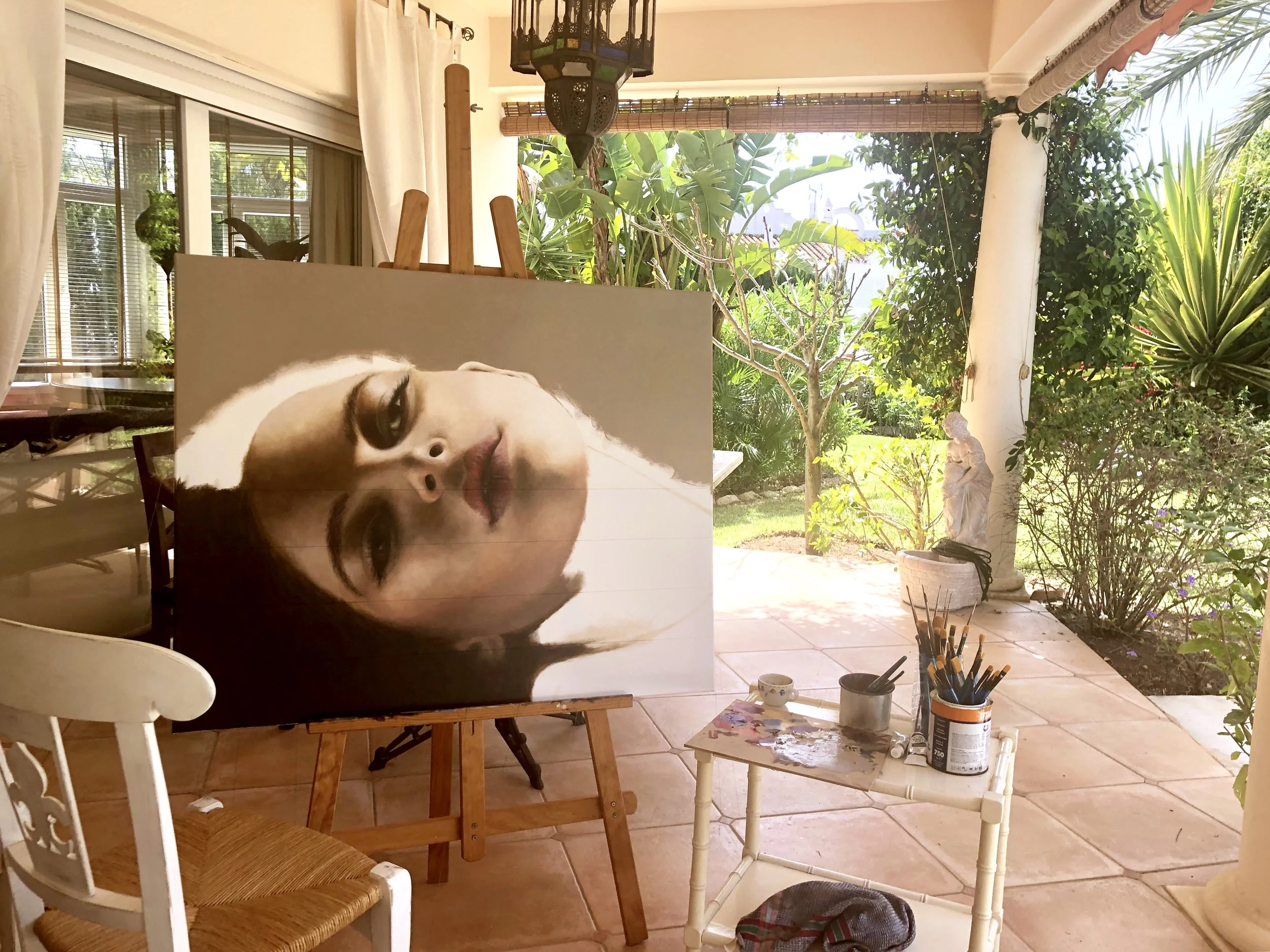 Indoor art studio with a large portrait of a woman's face on an easel, surrounded by plants outside and art supplies on a small table.
