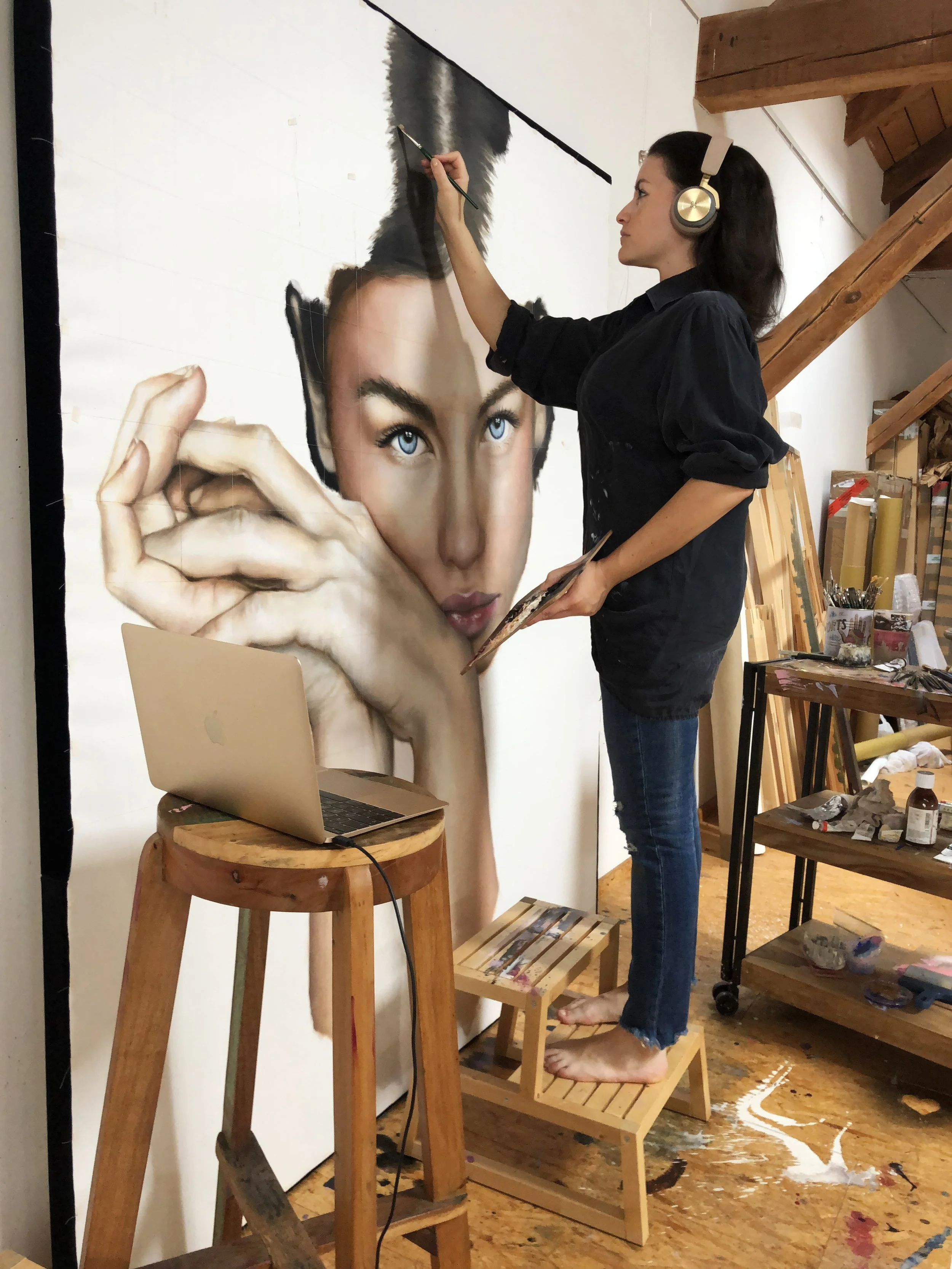 An artist is painting a large, realistic portrait of a woman with blue eyes and dark hair on a wall. She is standing on a small wooden step stool, wearing headphones, and holding a paint palette. There is a laptop on a wooden stool in front of her, with various art supplies and tools around the workspace.