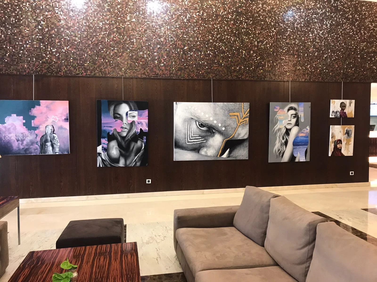 Gallery of five colorful paintings and three smaller artworks on a dark wooden wall in a modern lounge with beige sofa, dark ottoman, and a wooden table with green plant.