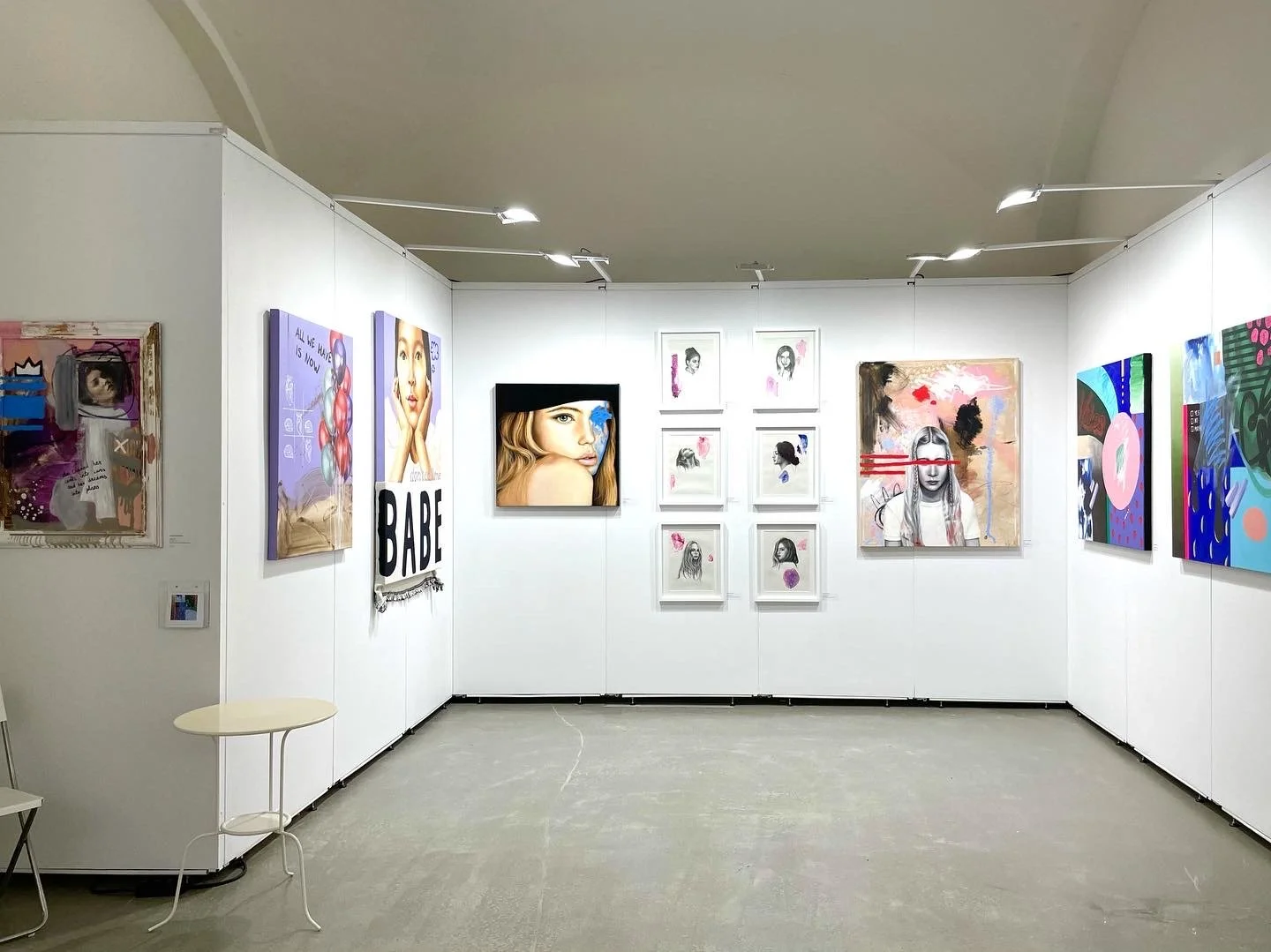 Art gallery with framed paintings and mixed-media artwork on white walls, featuring portraits and abstract designs, with ceiling track lighting.