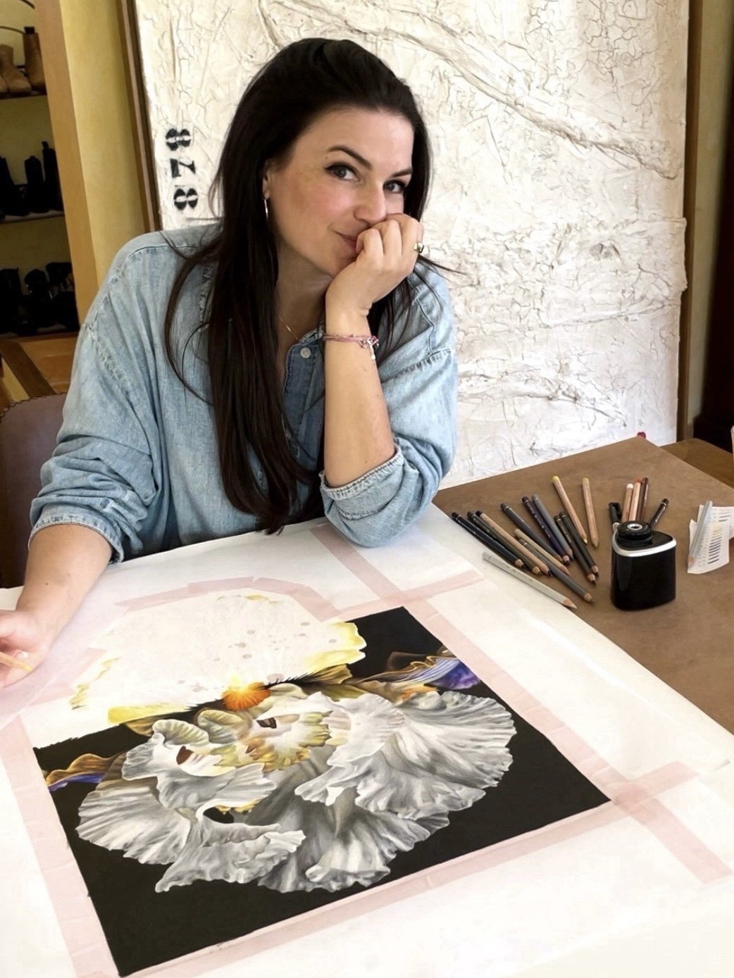 A woman with long dark hair, wearing a denim shirt, is sitting at a table with a detailed floral painting in progress. She is resting her chin on her hand, looking at the camera. Art supplies including colored pencils and markers are on the table.