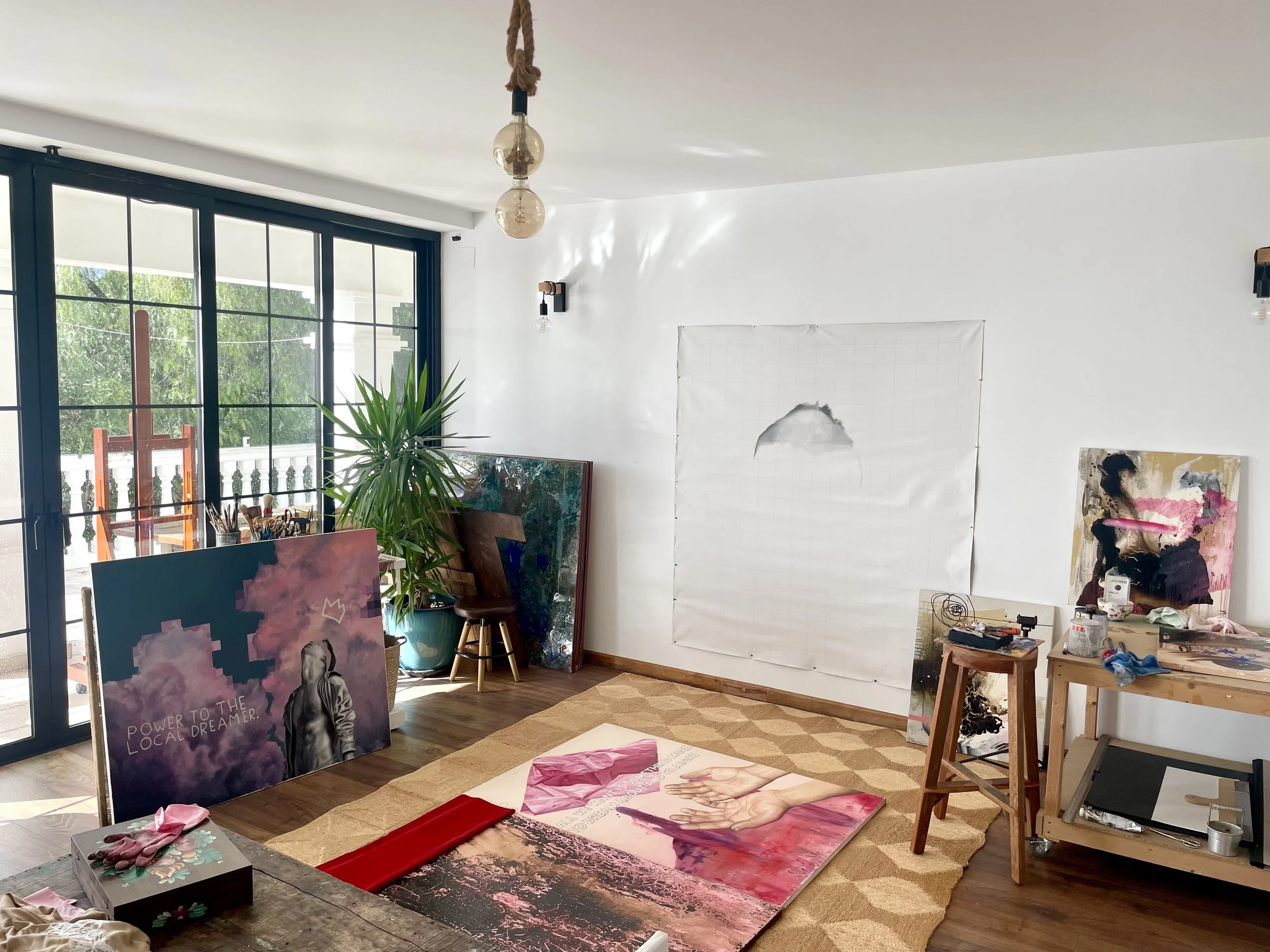 Artist's studio with large sliding glass doors, paintings, and art supplies. A painting on the floor features a pink and white theme with hands, and there are multiple other artworks leaning against the wall or standing. The room has wooden floors, a beige patterned rug, a wooden worktable, and a large potted plant near the window.