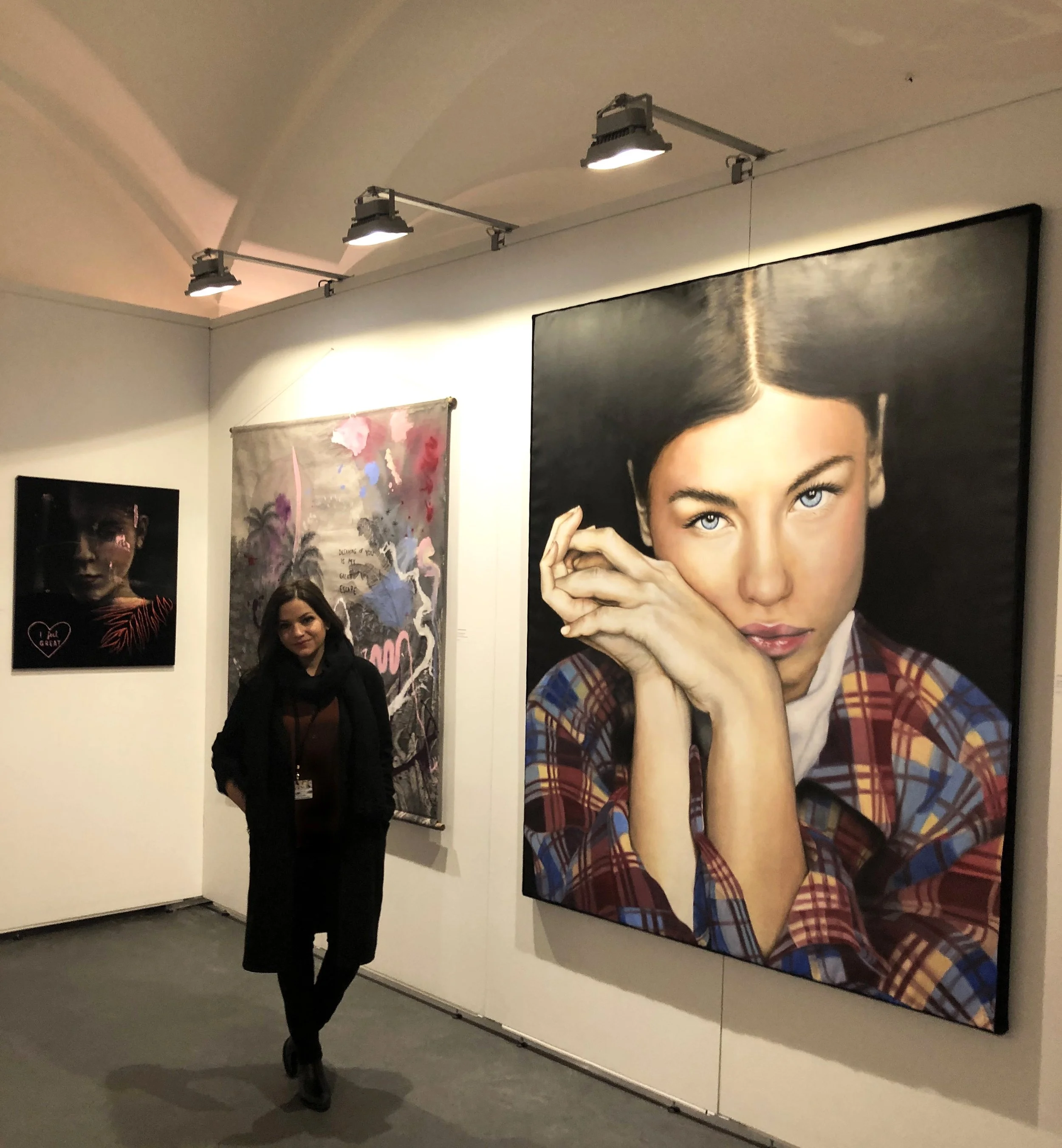 A woman standing in an art gallery next to a large portrait of a woman with blue eyes and dark hair, wearing a plaid shirt. There are other artworks displayed on the wall, and a woman is visible in the background.