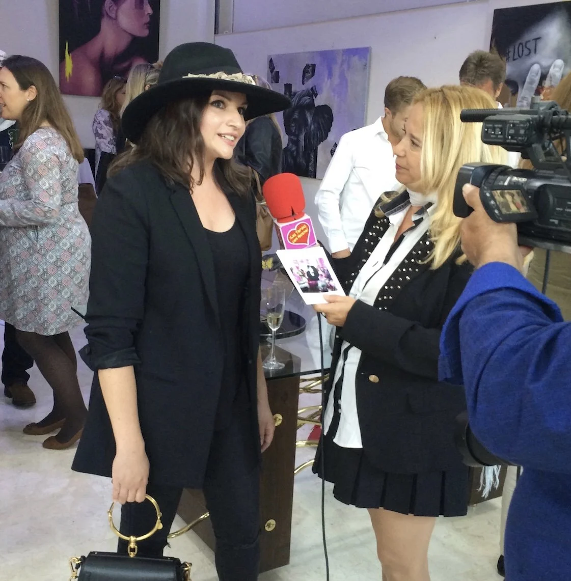 A woman wearing a black hat and black outfit is being interviewed at a social event, with a reporter holding a microphone and a camera operator filming. People are mingling and artwork is displayed on the walls in the background.
