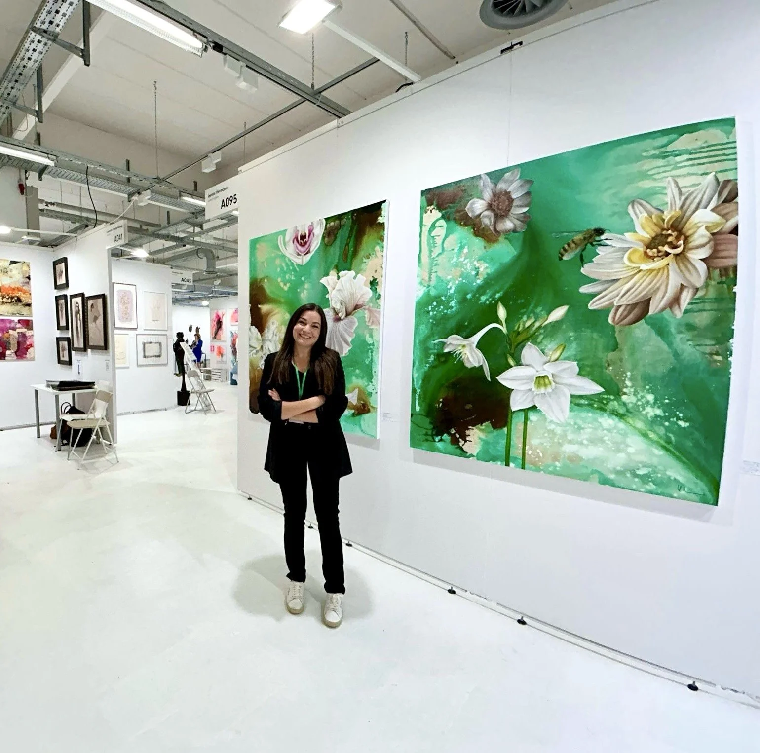 A woman standing in an art gallery with her arms crossed, smiling, next to two large paintings of white flowers and green backgrounds, surrounded by other artworks and gallery structures.