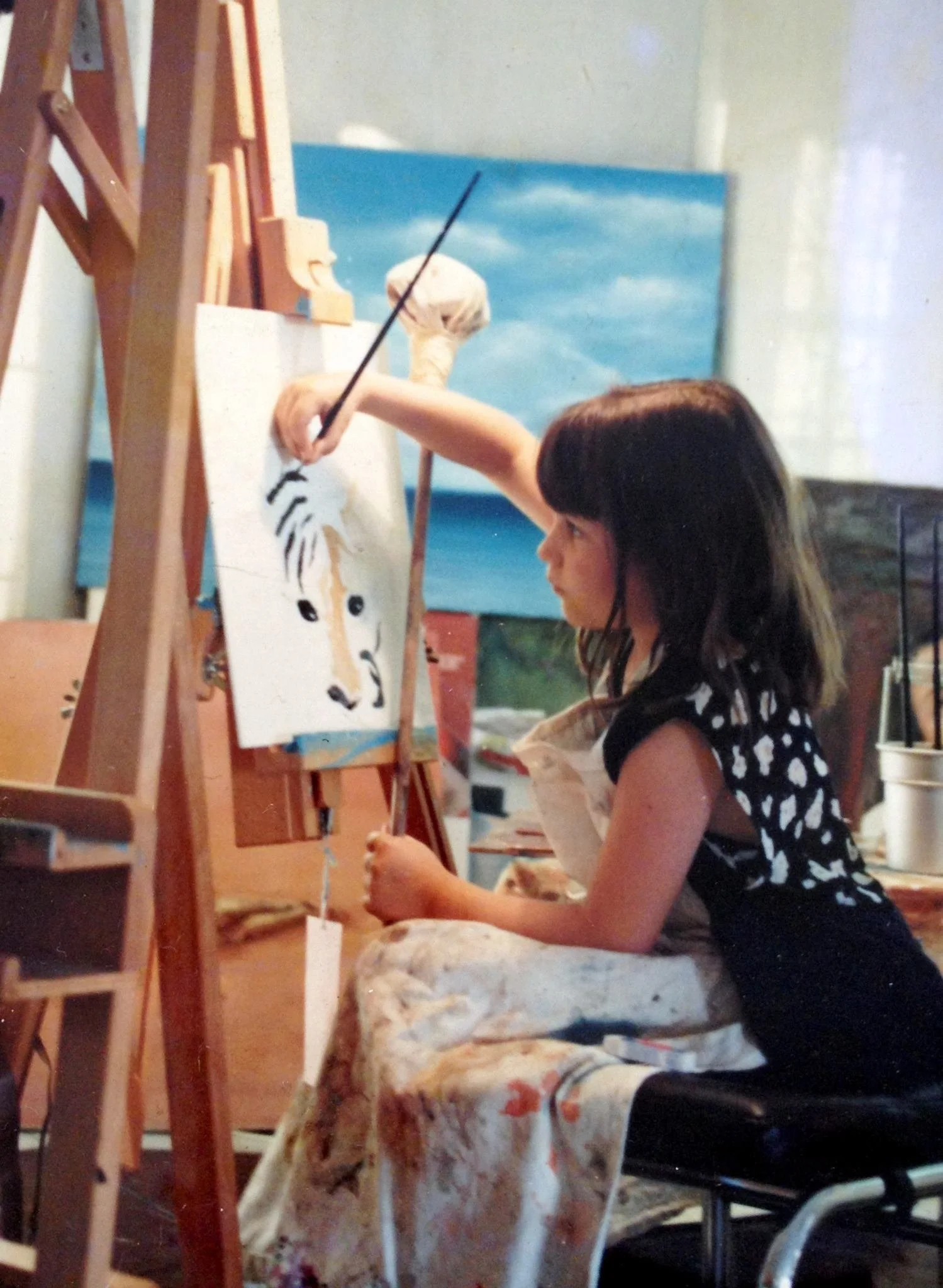 A young girl with dark hair painting a tiger on a canvas, seated in an art studio with paintings and art supplies around.