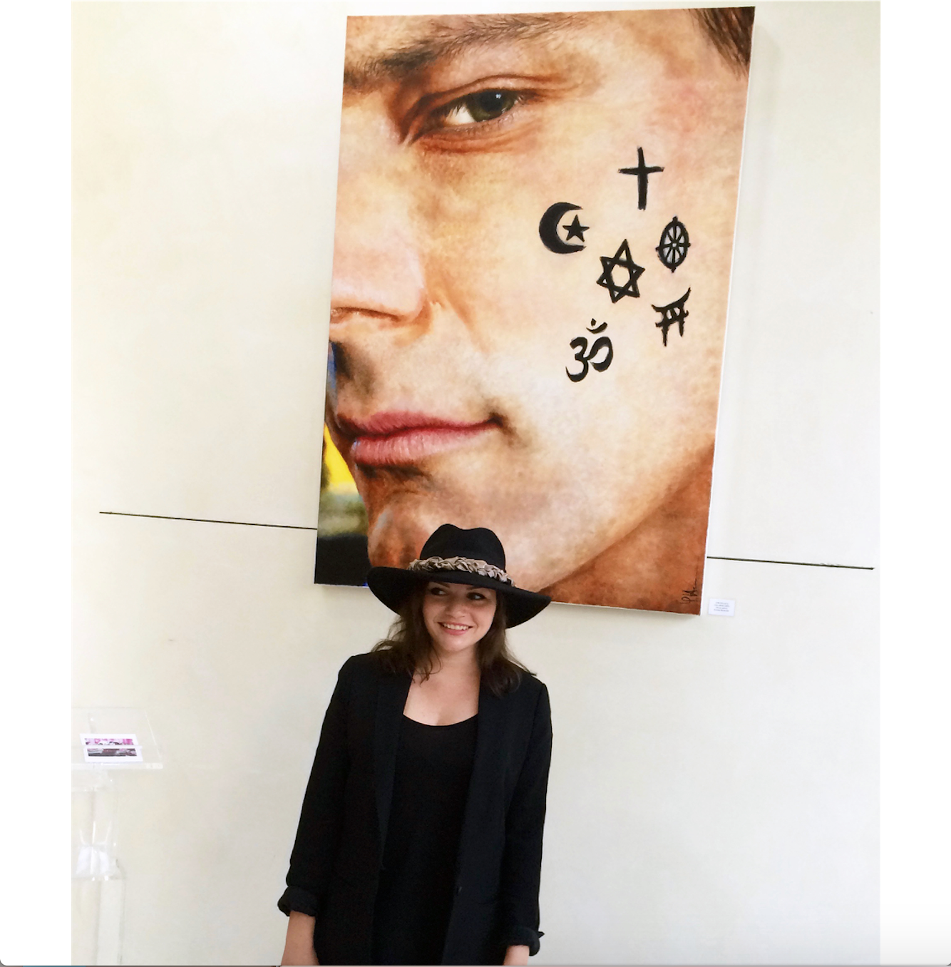 A smiling woman wearing a black hat and black blazer standing in front of a large artwork of a close-up of a man's face with various religious and spiritual symbols on his cheek.