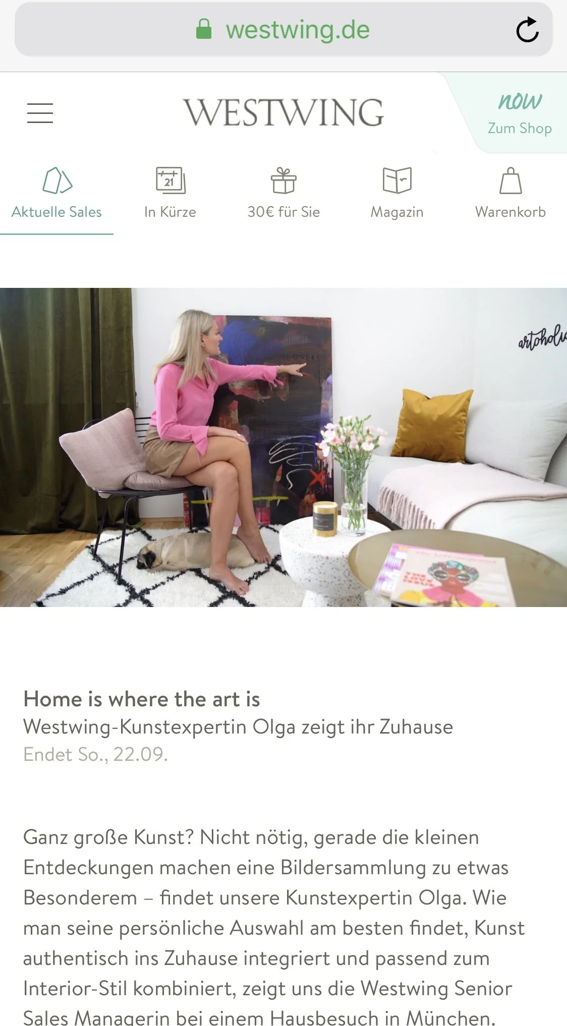 A woman sitting on a chair in a living room, pointing at an abstract painting. A pug lies on the floor beneath her. The room has a white sofa with yellow cushions, a round white coffee table with a flower vase and magazines, and dark green curtains.