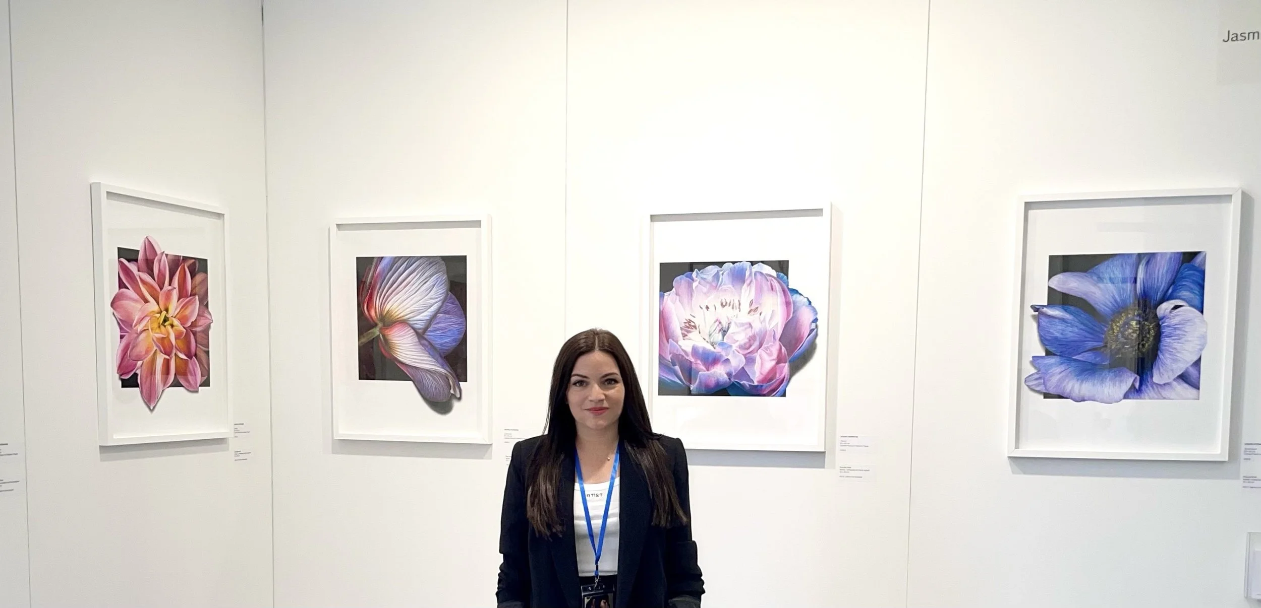 A woman with long dark hair and a blazer standing in front of framed floral artwork at an art gallery.