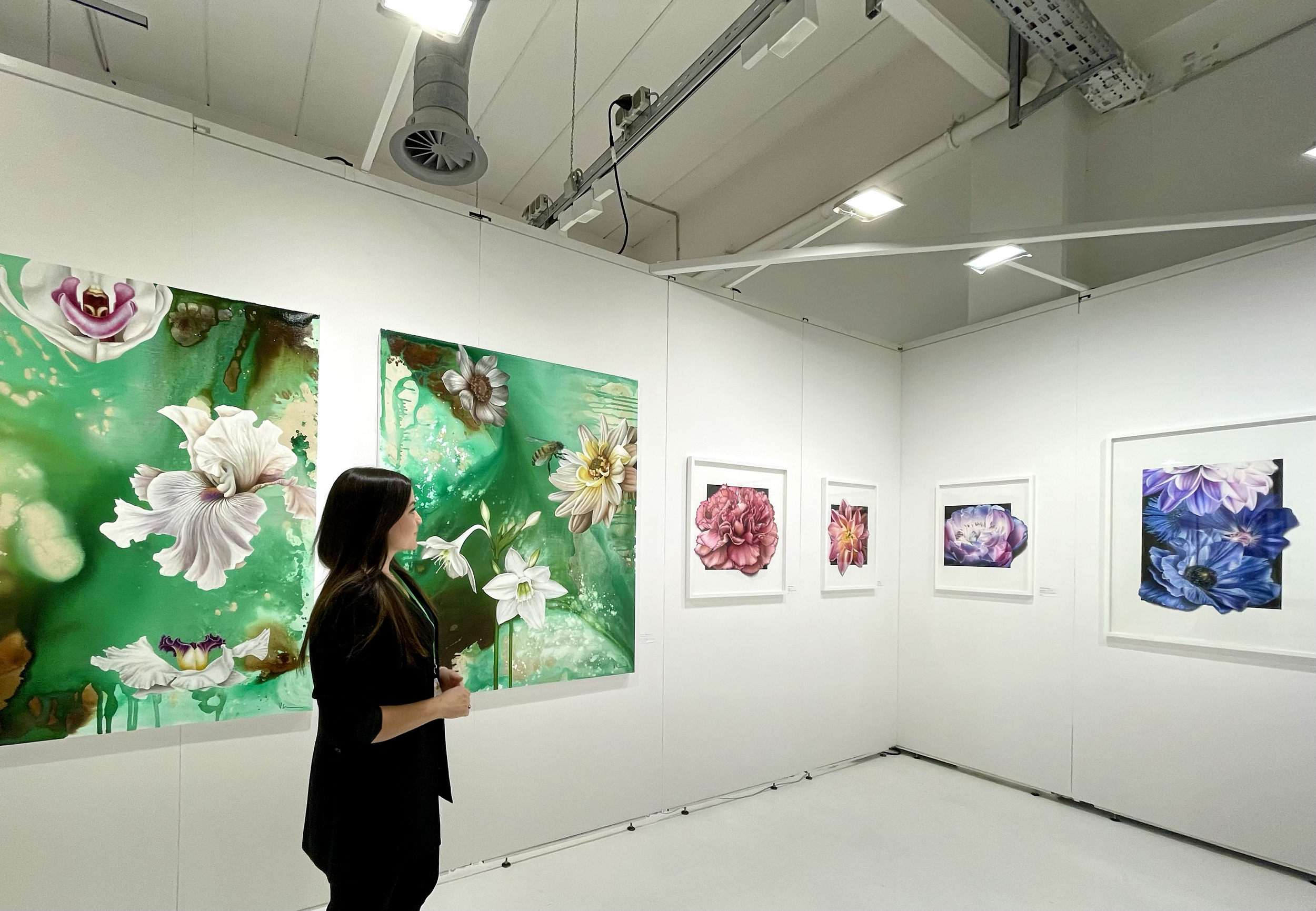 A woman stands in an art gallery looking at floral paintings on white walls. The artworks feature colorful, close-up depictions of flowers like lilies and peonies, with abstract backgrounds.