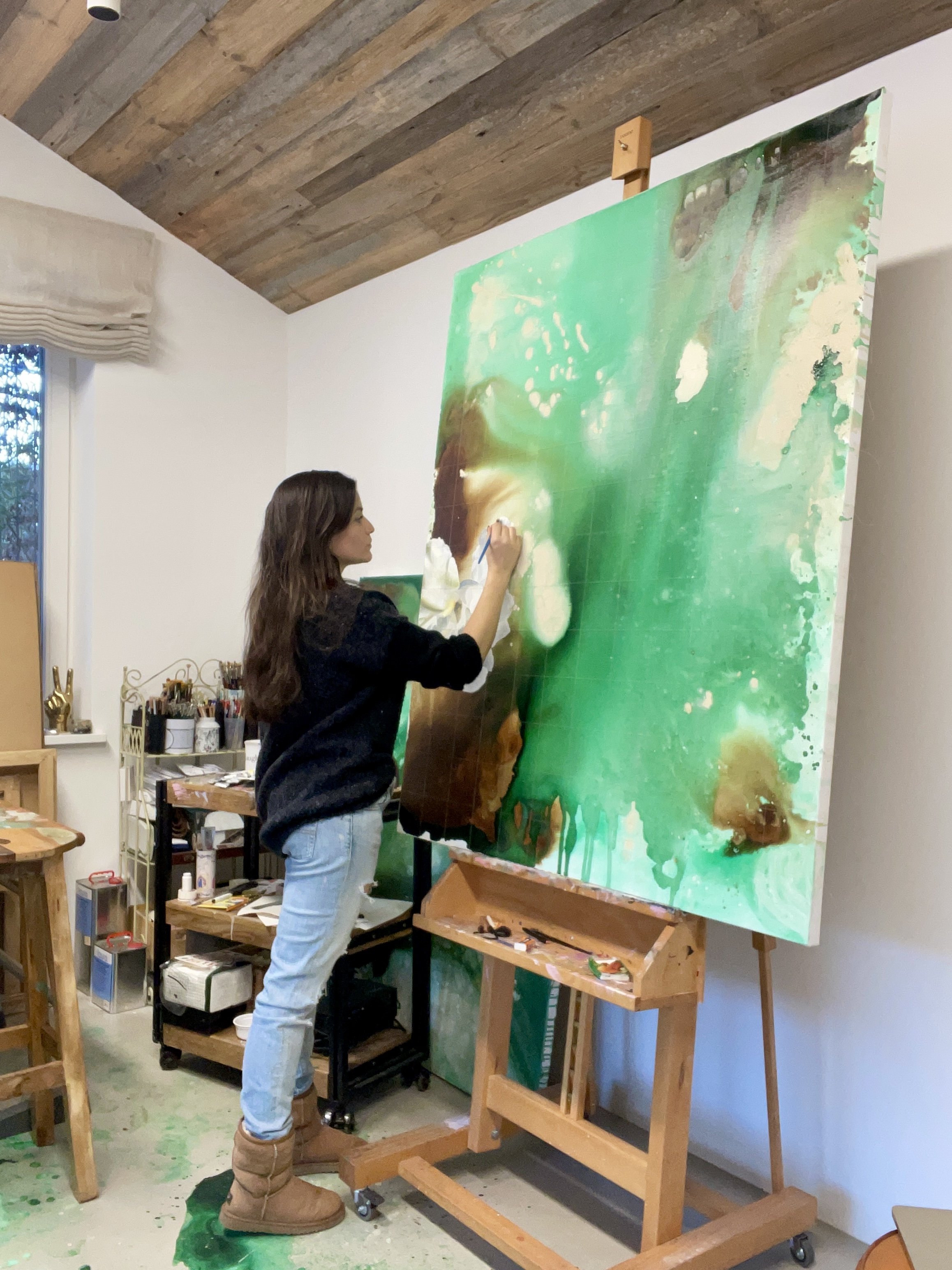A woman is painting an abstract artwork on a large canvas in a studio. The studio has wooden ceiling panels, a window, and art supplies