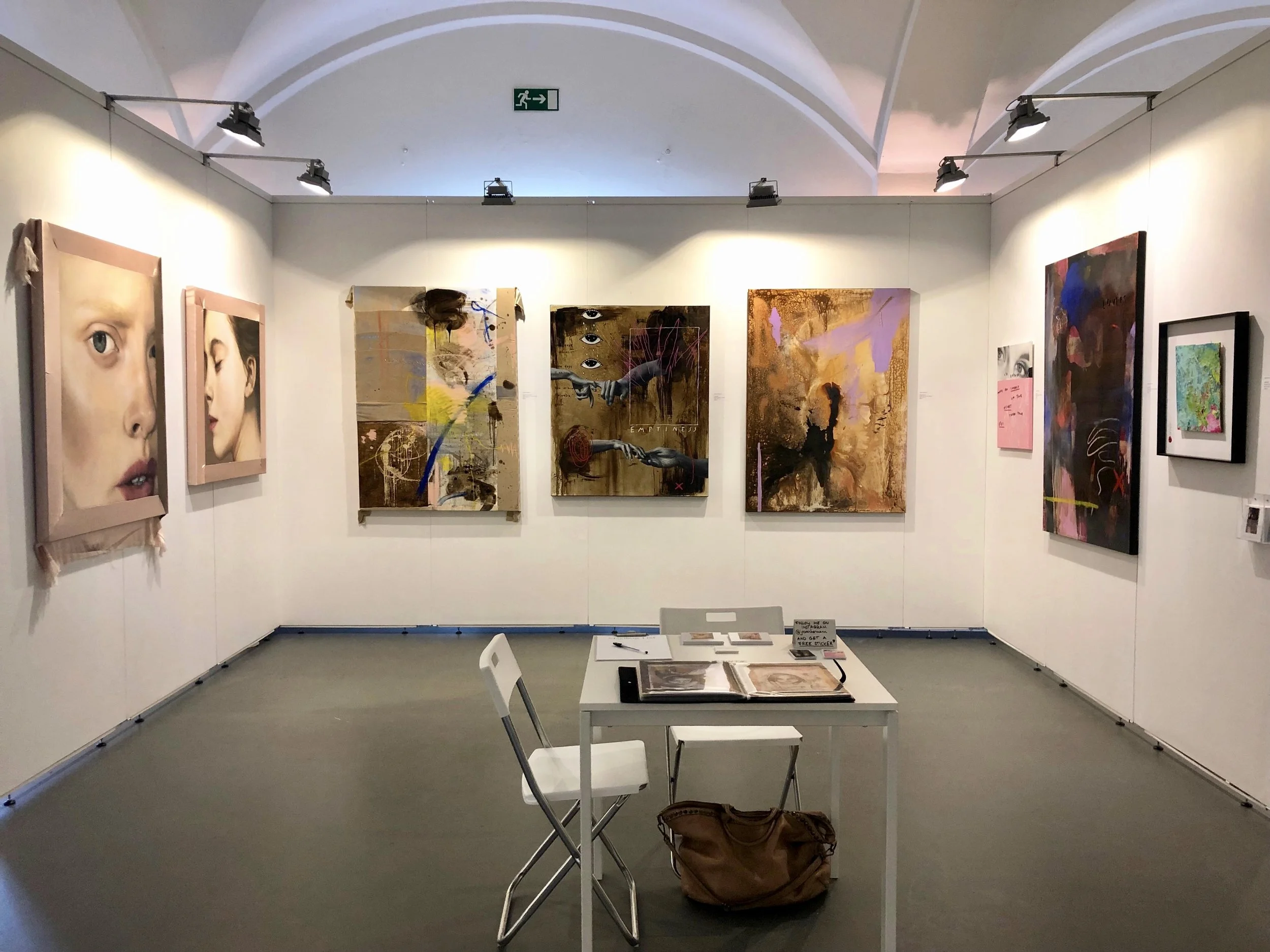 An art gallery with various contemporary paintings displayed on white walls. There is a small white table with chairs and art books in the center.