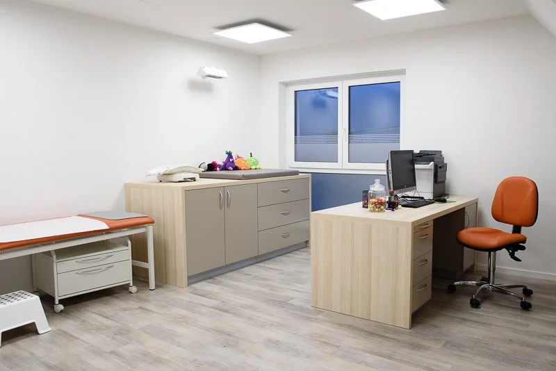 Innenansicht eines medizinischen Behandlungszimmers mit Untersuchungsbett, Schränken, Schreibtisch mit Computer und einem roten Bürostuhl, Fenster mit blauer Sicht, dezent beleuchtet.