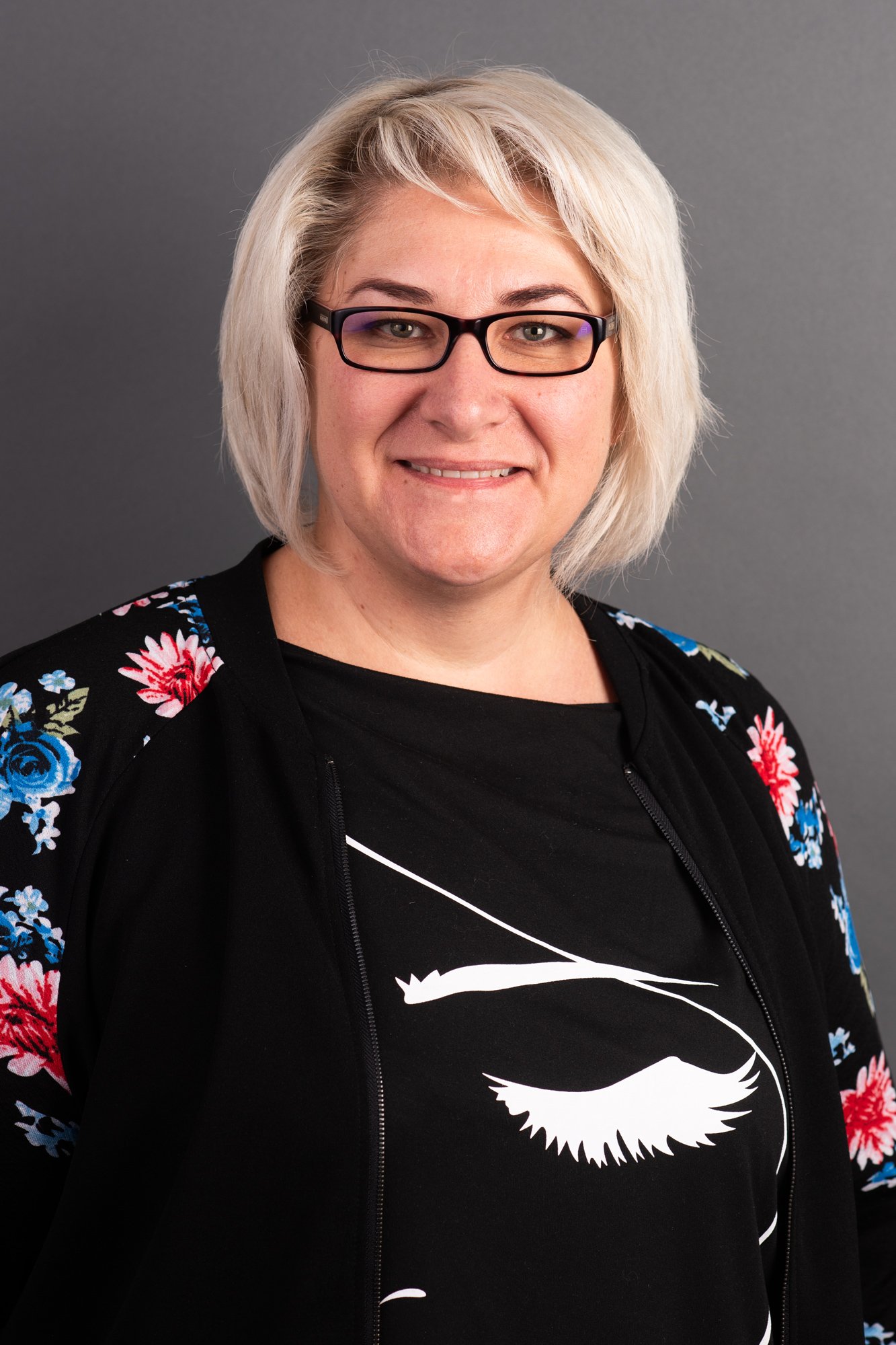 Porträt einer Frau mit Brille und blonder, kurz gewellter Frisur, trägt ein schwarzes T-Shirt mit Stierkopf-Logo und eine Jacke mit Blumendruck, vor grauem Hintergrund.