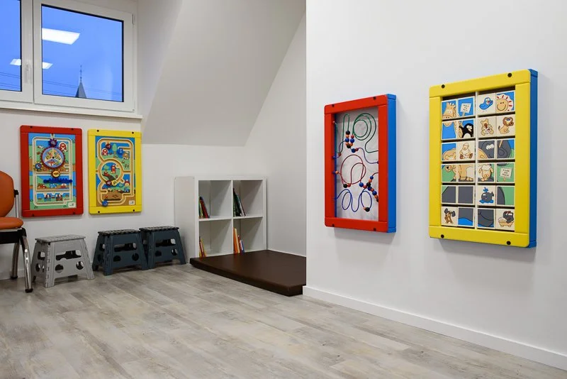 Ein ruhiger Wartebereich oder Spielraum für Kinder mit bunten Spieltafeln an den Wänden, einem kleinen Regal mit Büchern, drei Stapelhockern, einem Stuhl und einem Sitzkissen, unter einem Fenster mit Blick nach draußen.