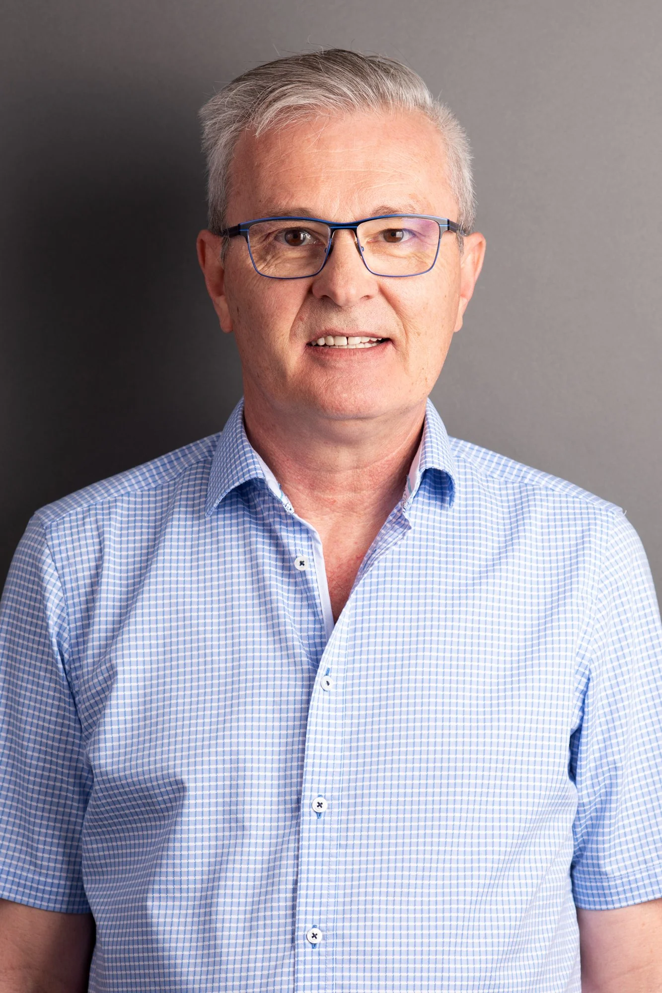 Ein Mann mit grauem Haar, Brille und blau-weiß kariertem Hemd vor grauem Hintergrund.