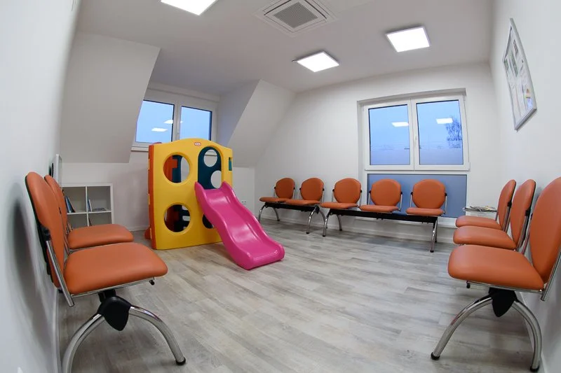 Warteraum mit orangefarbenen Stühlen, Kinder-Klettergerüst mit Rutsche, Fenster und Deckenleuchten.