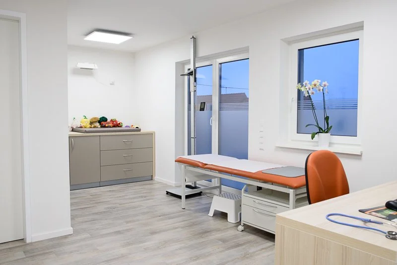 Behandlungsraum in einer Arztpraxis mit Untersuchungsliege, Fenster, Orchidee und Schrank mit medizinischem Zubehör.