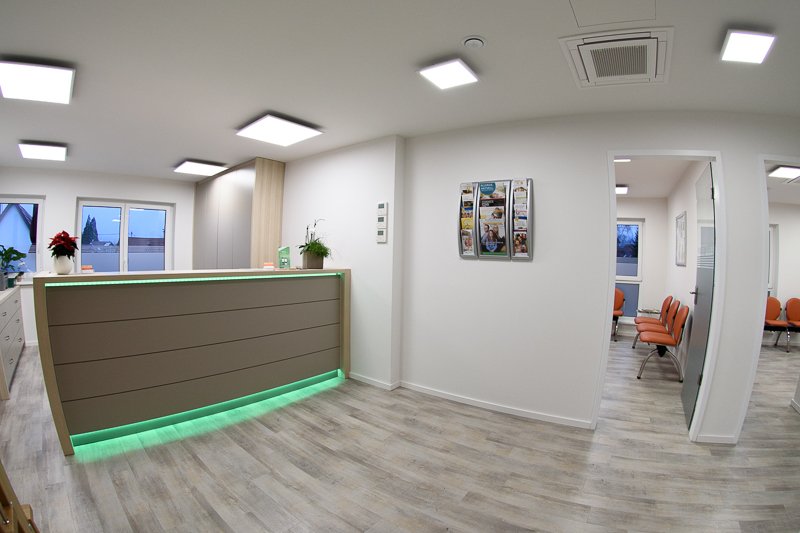 Wartezimmer mit Empfangstheke, Stühlen und Informationsbroschüren auf der Wand. Helle, moderne Einrichtung mit Deckenleuchten.