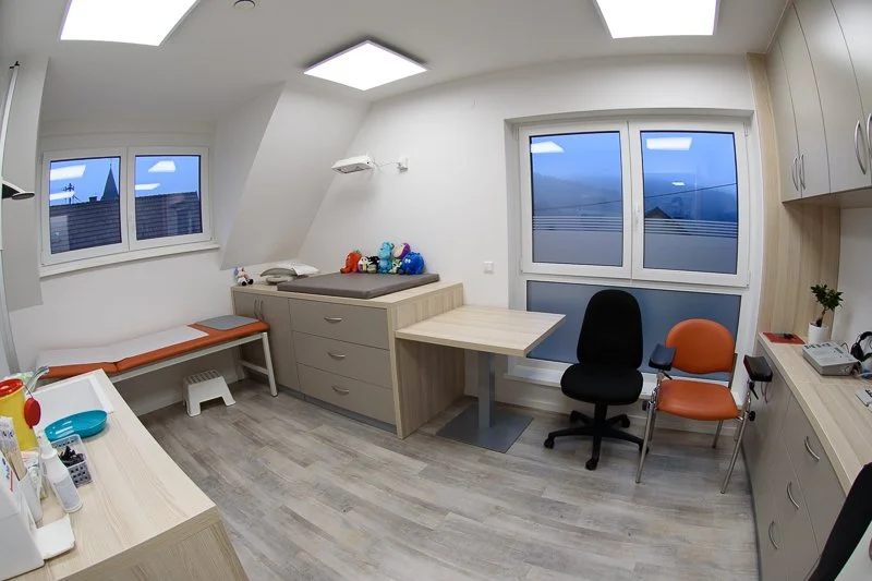 Ein Klinik- oder Behandlungszimmer mit Fenstern, einem Untersuchungstisch, Schränken, einem Schreibtisch und Stühlen.