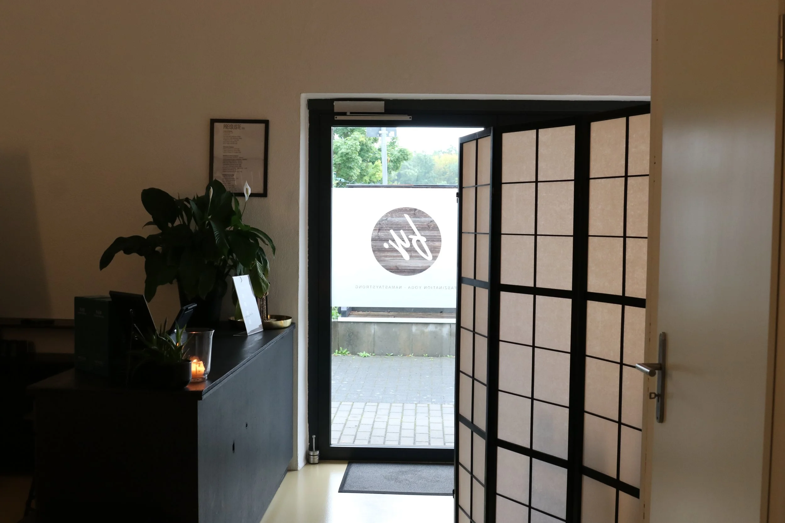 Innenansicht eines Eingangsbereichs mit einer schwarzen Konsole, einer Pflanze, einer Kerze und einer schwarzen Raumtrennwand, die einen Blick nach draußen auf eine Straße mit Bäumen erlaubt.