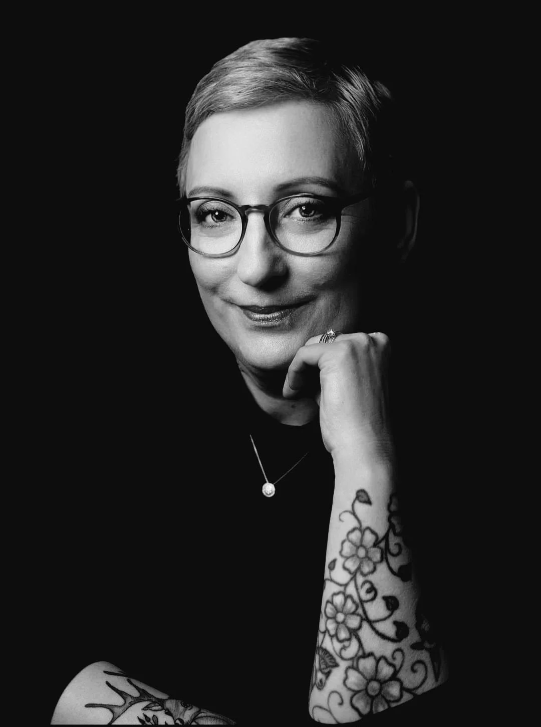 Schwarz-weiß-Porträt einer Frau mit kurzen, glatten Haaren, Brille und Tattoo auf dem Arm, die ihre Hand an das Kinn legt.