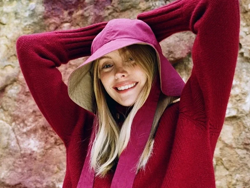 A smiling woman wearing a maroon bucket hat and matching sweater stands against a rocky background, with her hands behind her head.