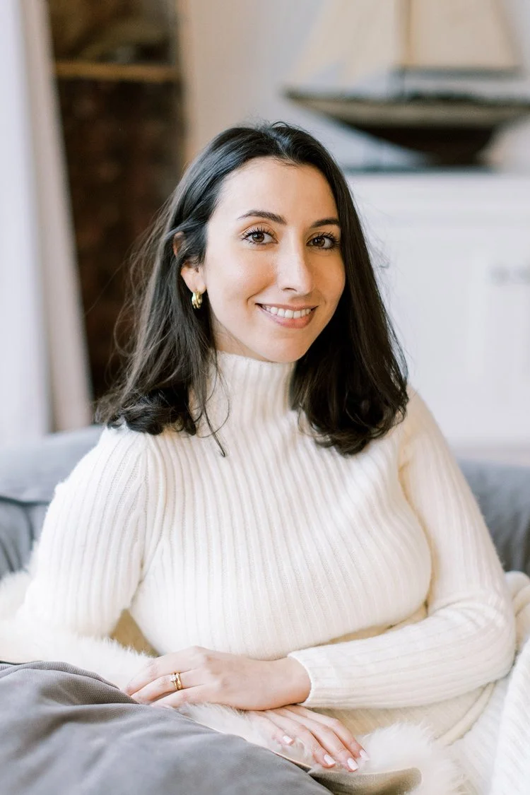 Photo of Victoria Locke, who has long dark hair, wearing a white ribbed turtleneck dress, sitting on a couch with a smile.
