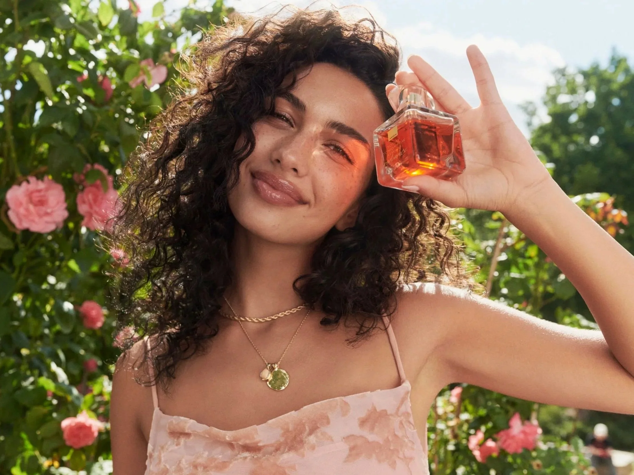 A young woman with curly hair in a pink dress holding a perfume bottle outdoors among pink roses and green foliage, smiling.