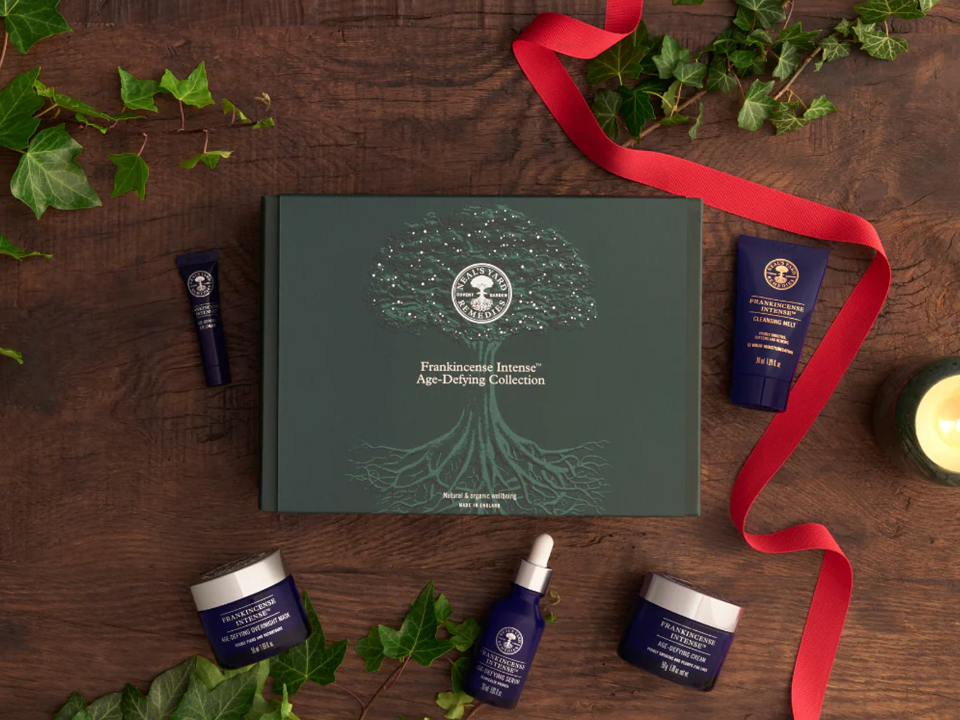 A collection of skincare products from the Frankincense Intense Age-Defying Collection arranged on a wooden surface around Christmas packaging with green ivy leaves and a red ribbon surrounding them, and a candle in a jar to the right.