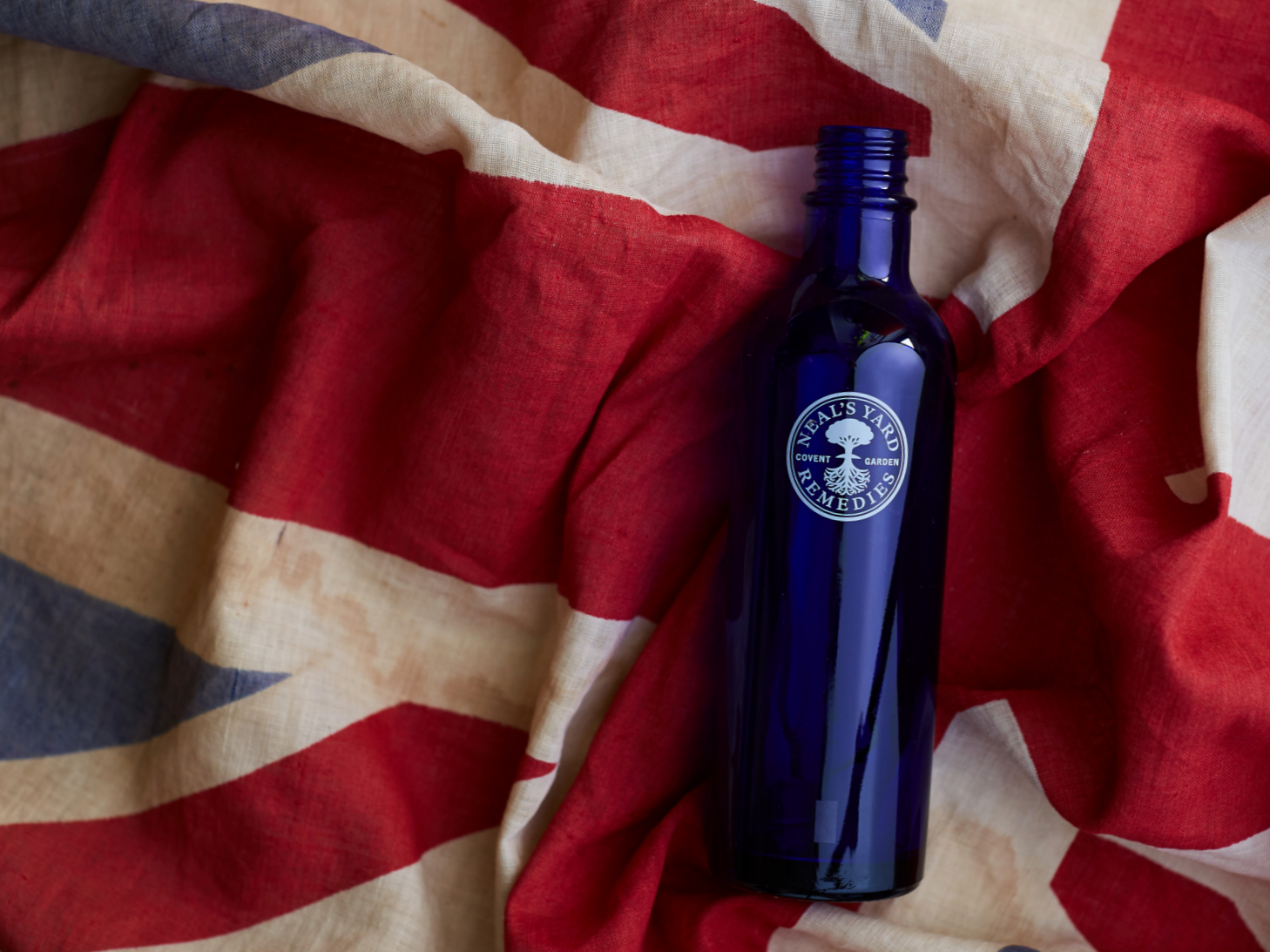 Blue glass bottle with a label reading 'Neal's Yard Remedies' placed on a crumpled fabric featuring the UK flag.