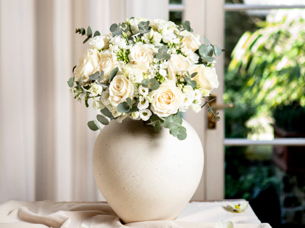 A large cream-colored vase with a bouquet of white roses, small white flowers and eucalyptus leaves on a table near a window.