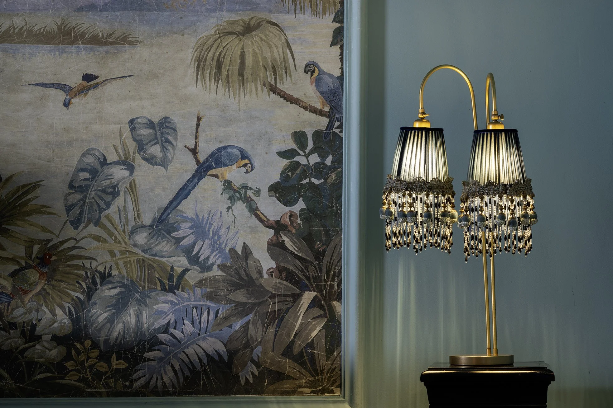 A decorative painting of tropical birds and lush foliage on the wall, next to a lamp with a black base and a double lampshade with dangling beads and jewelry embellishments.