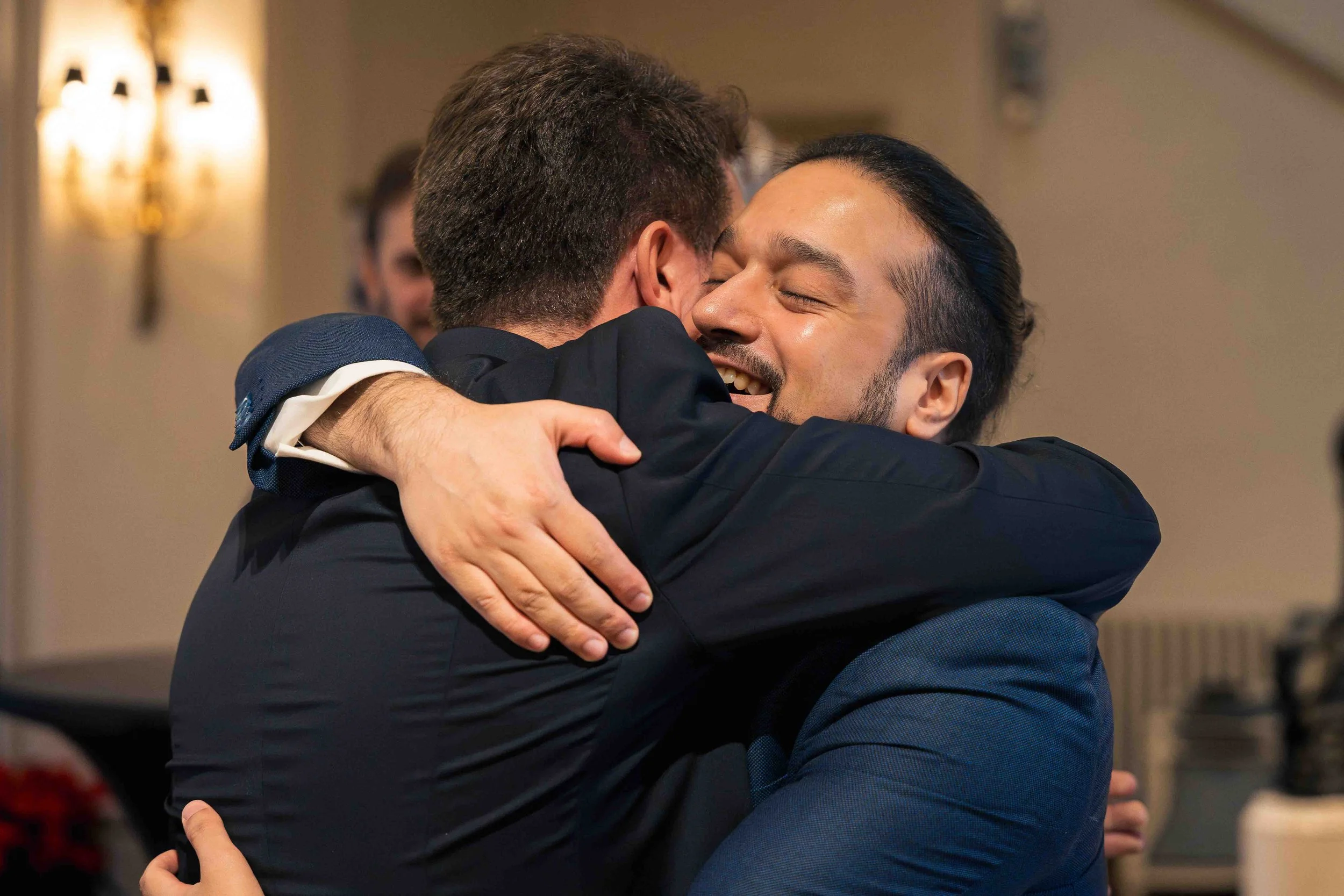 Photographie documentaire capturant un moment d’émotion lors d’un événement social ou familial. Deux hommes en costume s’enlacent chaleureusement dans une étreinte, exprimant la joie et la complicité. L’arrière-plan légèrement flou et la lumière inté