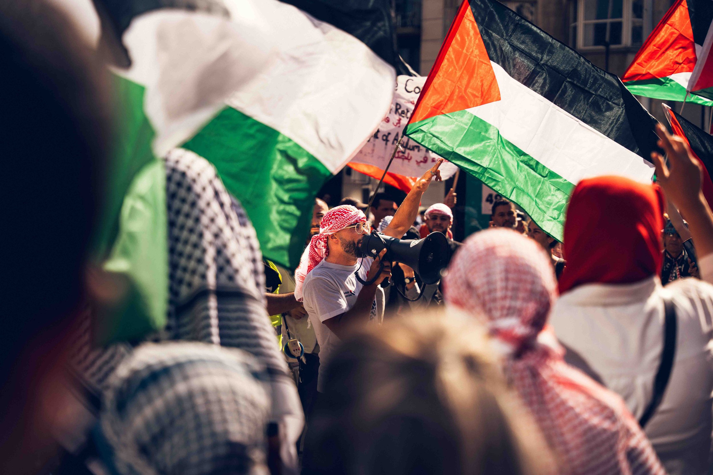 Photographie documentaire réalisée lors d’une manifestation pro-palestinienne dans l’espace public. Un manifestant s’exprime au mégaphone au milieu d’une foule brandissant des drapeaux palestiniens. L’image capture l’intensité du rassemblement, les s