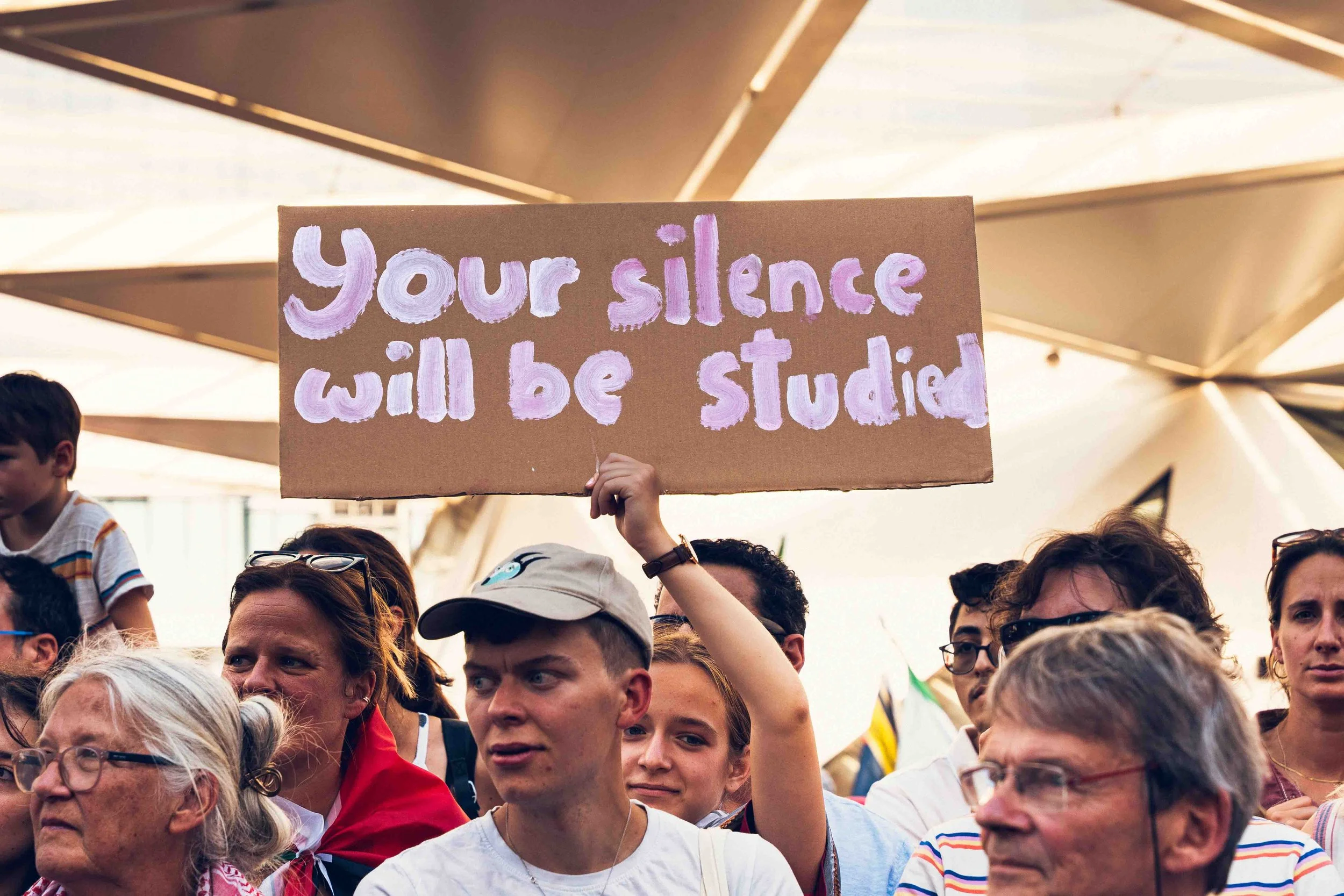 Photographie de reportage capturant des participants lors d’une manifestation dans l’espace public. Au centre de l’image, une personne tient une pancarte portant le message « Your silence will be studied », entourée d’une foule rassemblée. L’image do