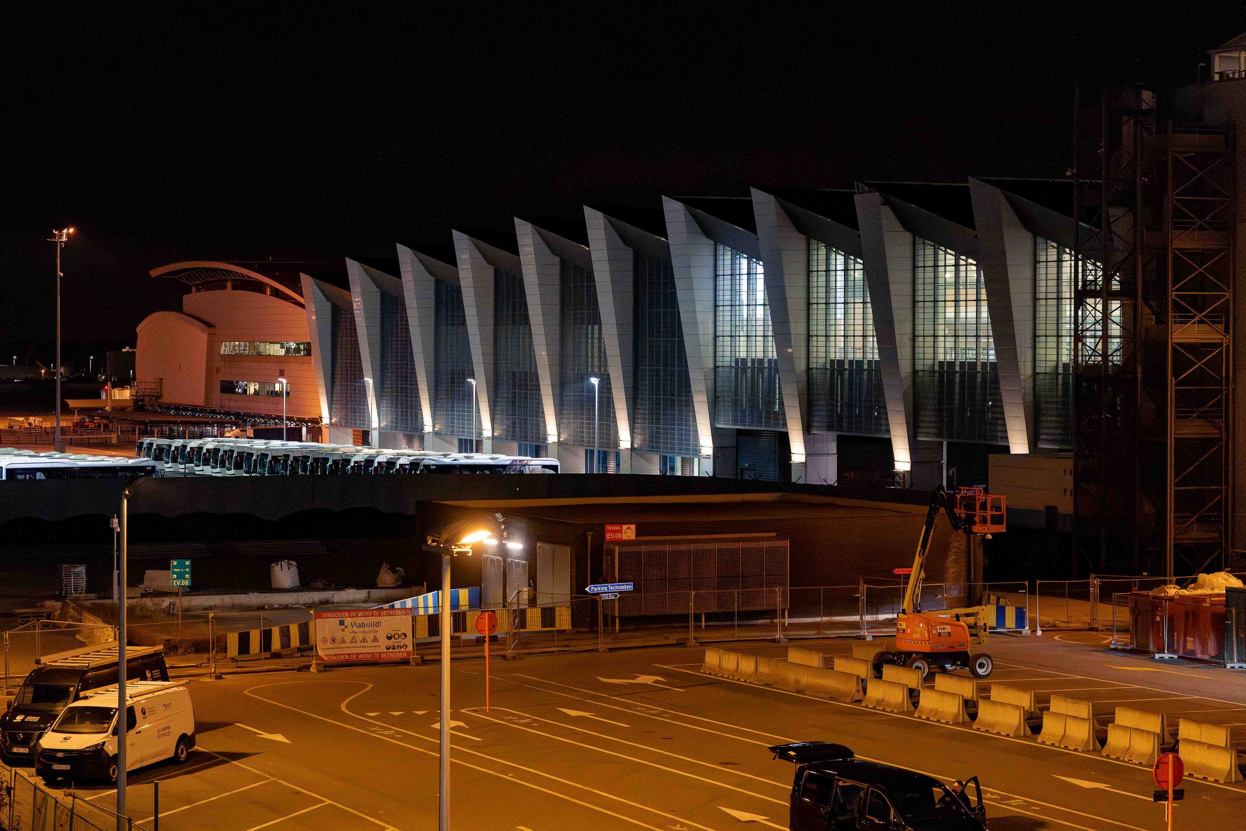 Photographie nocturne d’un bâtiment contemporain aux lignes architecturales marquées, illuminé par des éclairages extérieurs. La façade composée de structures répétitives et de grandes surfaces vitrées se détache dans la nuit, tandis que la zone urba